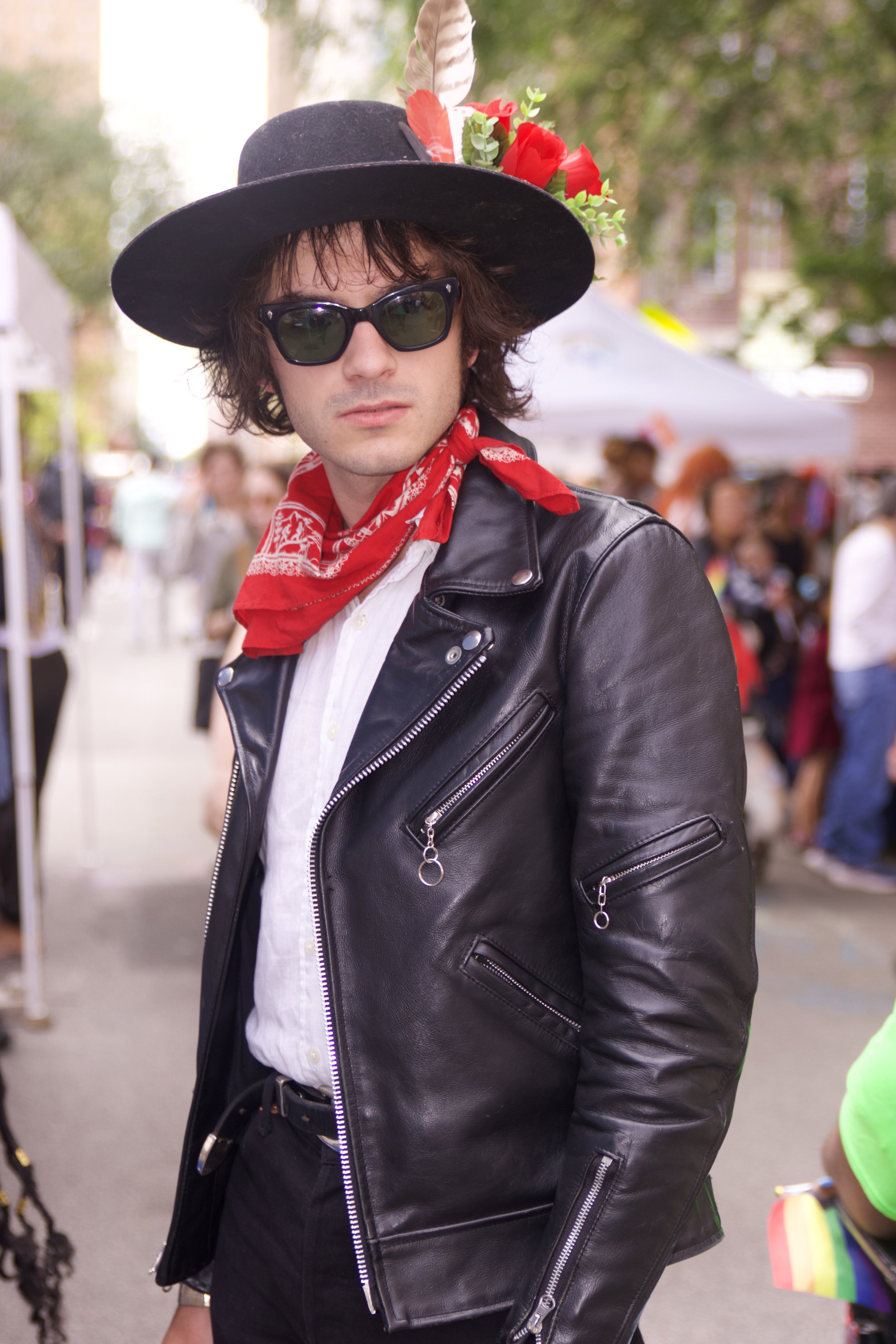 A man with long dark hair wearing black sunglasses, a black wide-brimmed hat decorated with a white feather and red flowers, a red bandana around his neck, a white shirt, and a black leather jacket, standing outdoors at what appears to be a festival or market with blurred people and tents in the background.