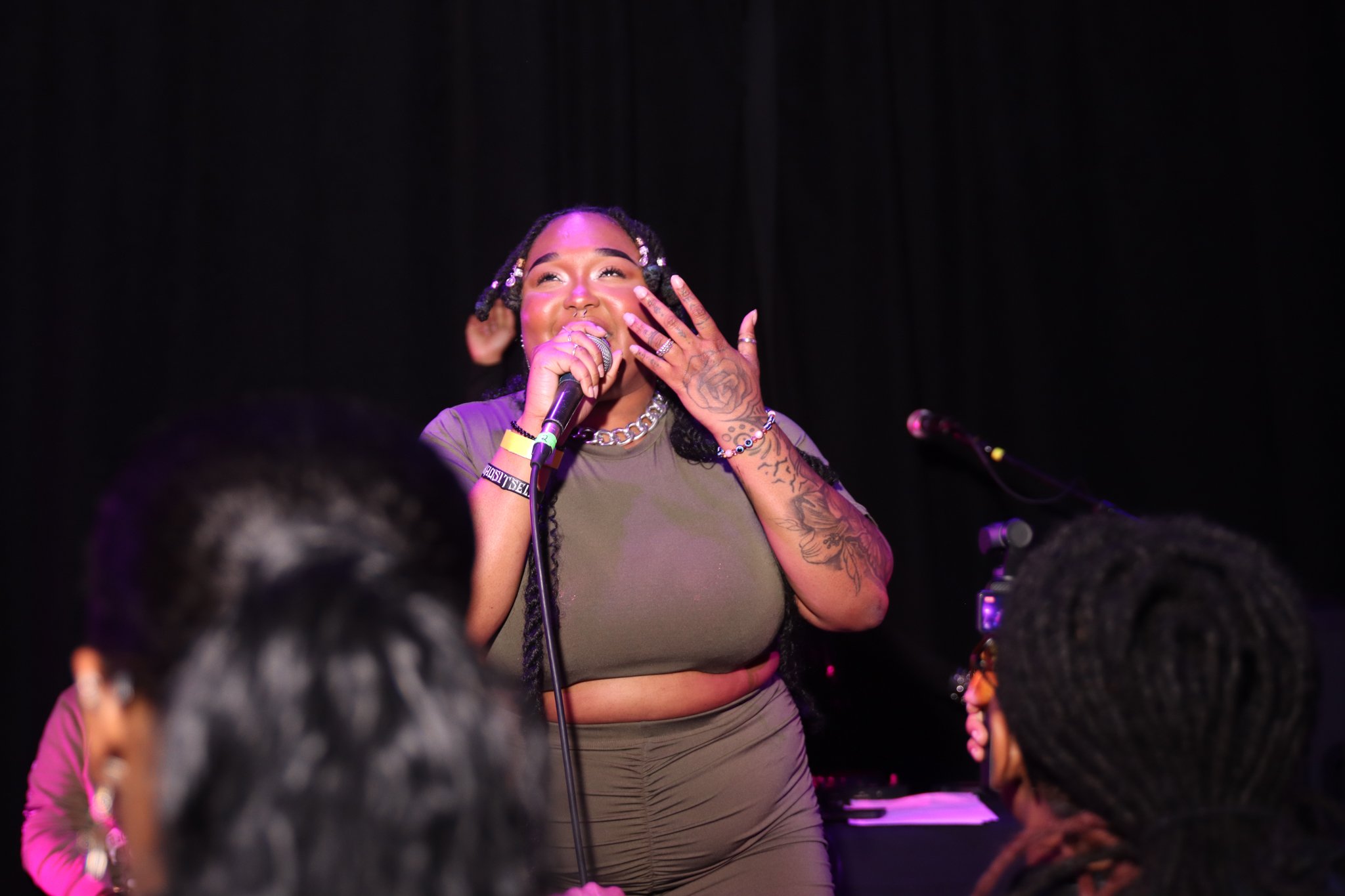 A woman singing on stage with a microphone, wearing a green top and matching pants, with multiple tattoos on her arms and jewelry, and engaging with the audience.