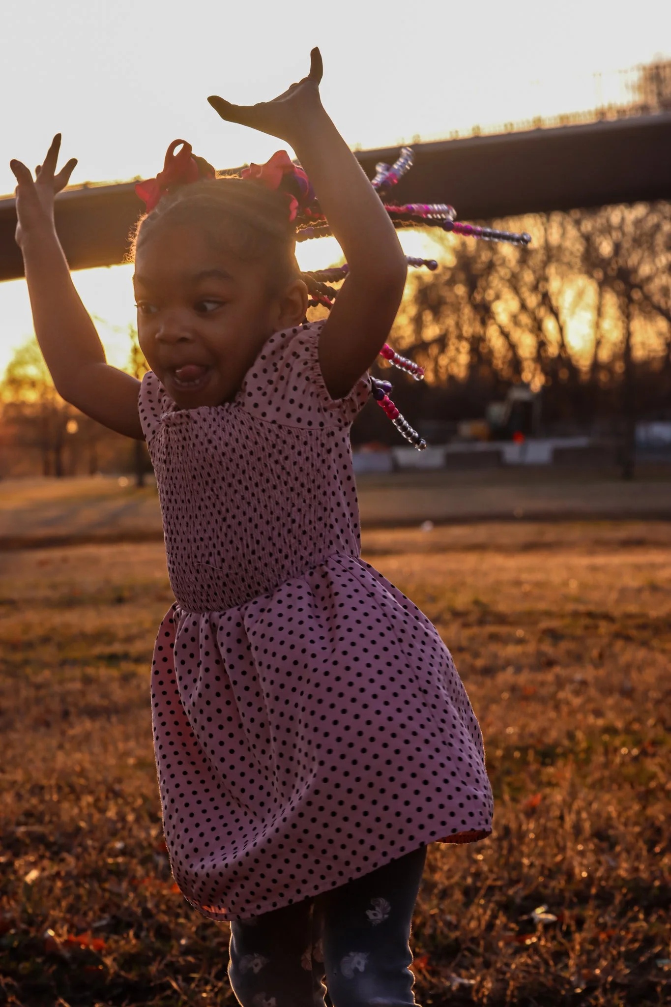 A young girl with beads in her hair, wearing a pink polka dot dress, playing outside during sunset.