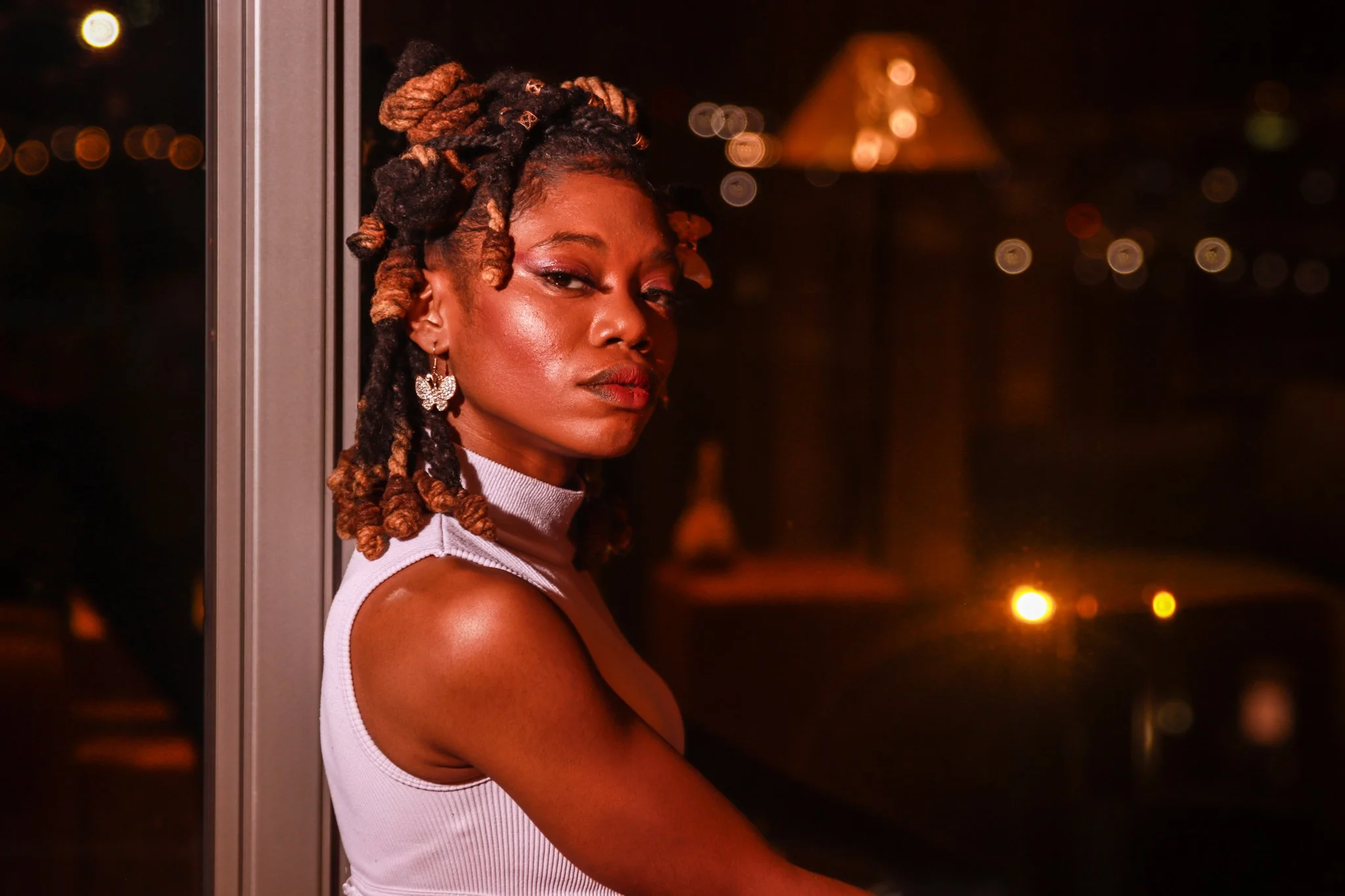A woman with styled dreadlocks wearing a light-colored sleeveless top and butterfly-shaped earrings, looking over her shoulder at the camera, with blurred city lights in the background at night.