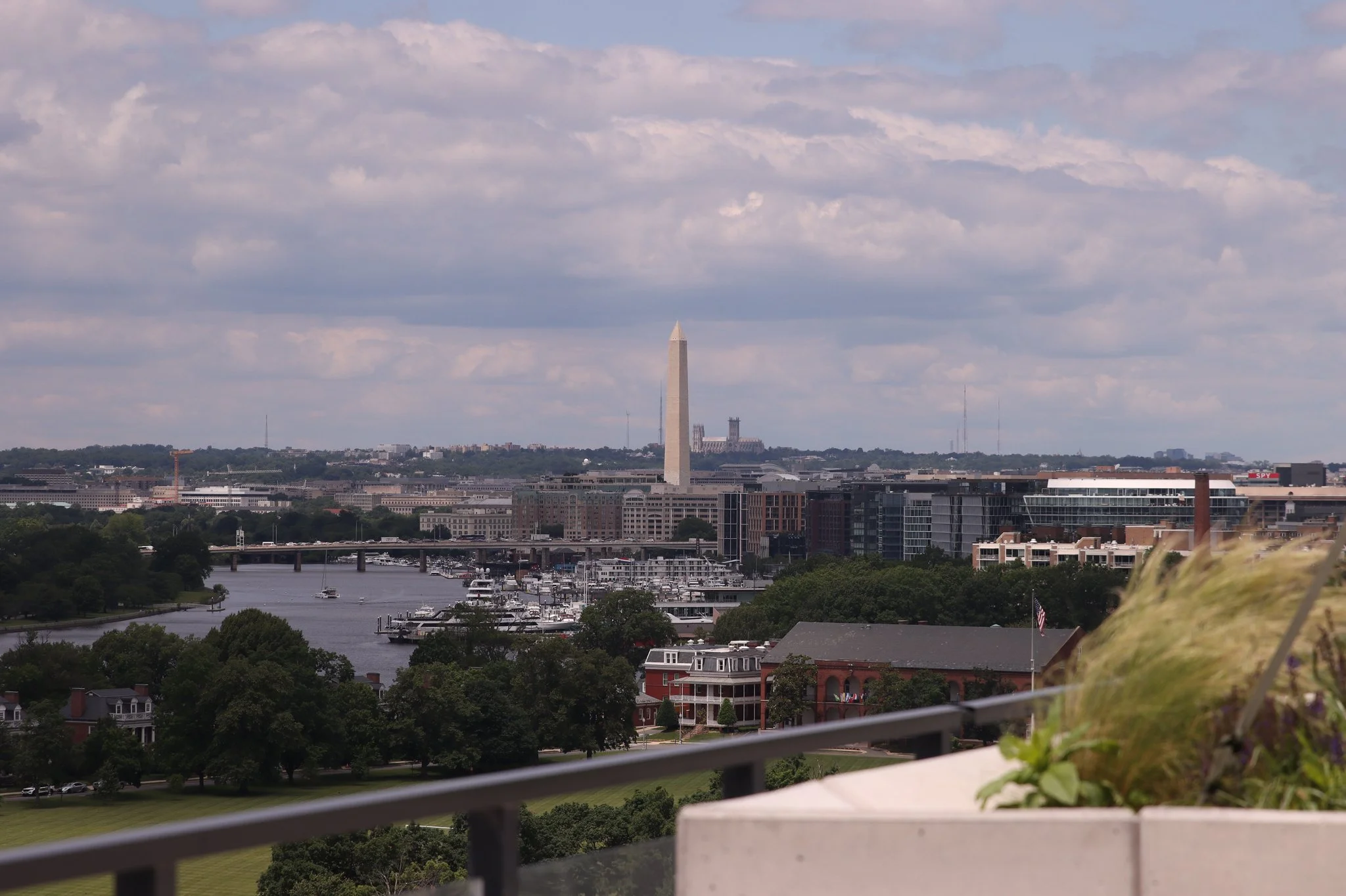 View of Washington, D.C., featuring the Washington Monument, the U.S. Capitol building in the distance, and a river with boats, under a partly cloudy sky.