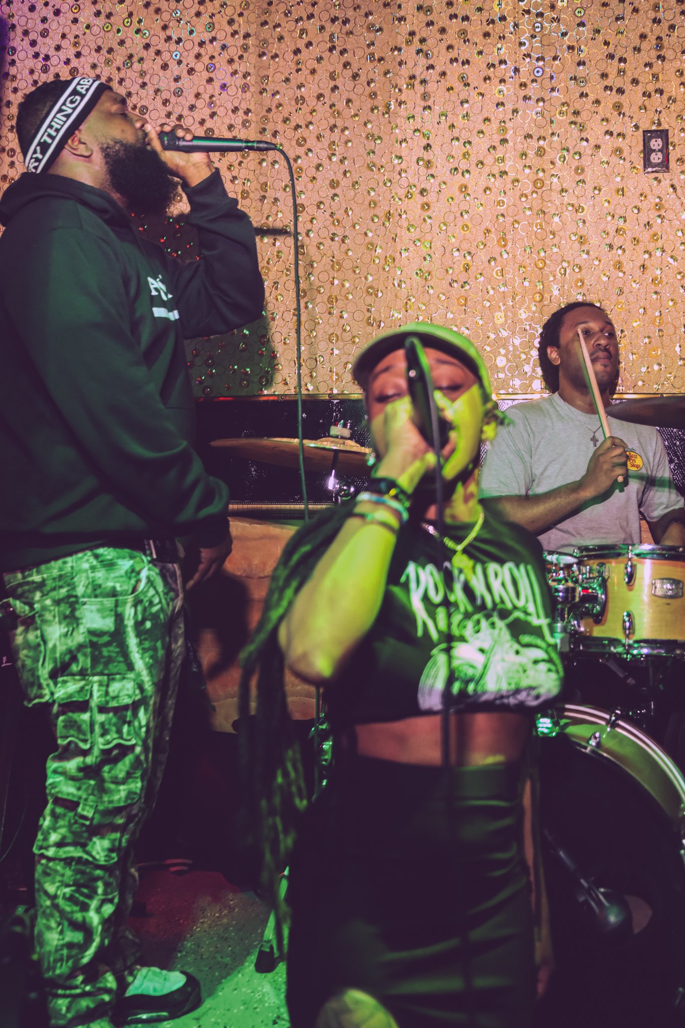 Three musicians performing on stage with a decorated gold wall in the background. The man on the left is singing into a microphone, wearing a black hoodie and camouflage pants. The woman in the center is singing into a microphone, wearing a black crop top and headphones. The man on the right is playing drums, wearing a gray t-shirt.