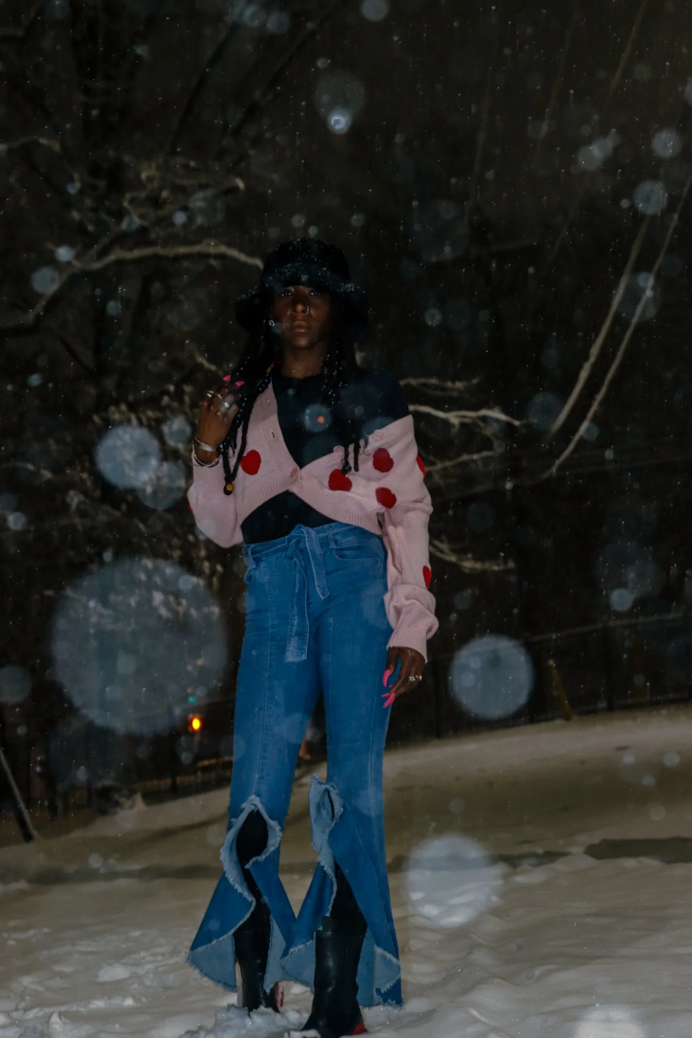 A woman standing on snow at night, wearing a pink cardigan with red polka dots, blue ripped jeans, black boots, and a black hat, with snow falling around her.