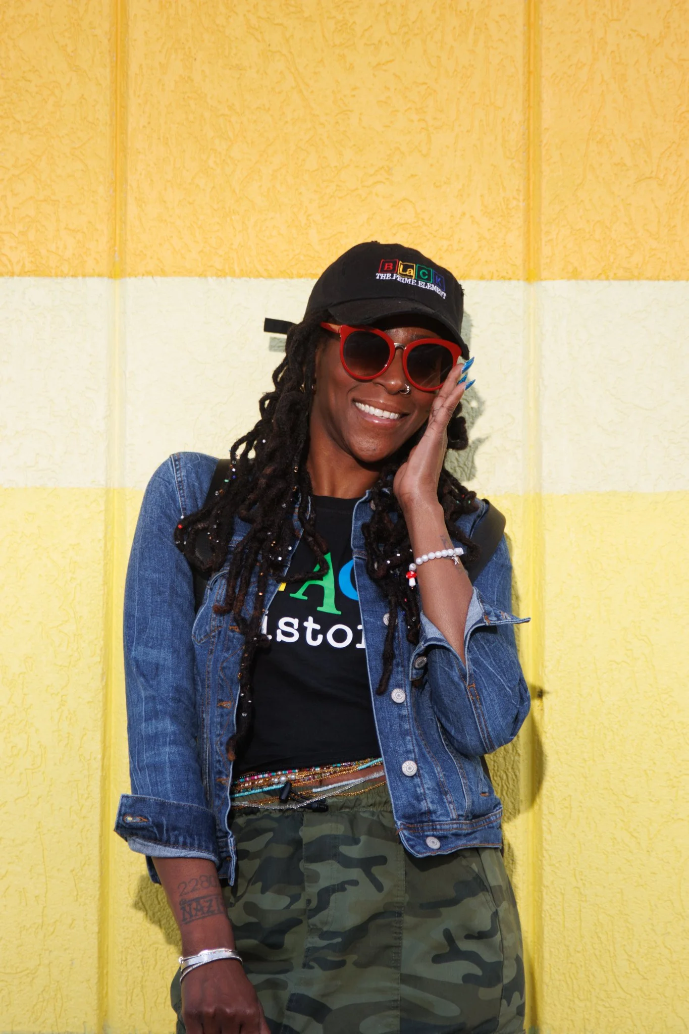 Woman smiling, wearing red sunglasses, a black cap, a denim jacket, camouflage pants, and colorful beaded accessories, standing against a yellow textured wall.