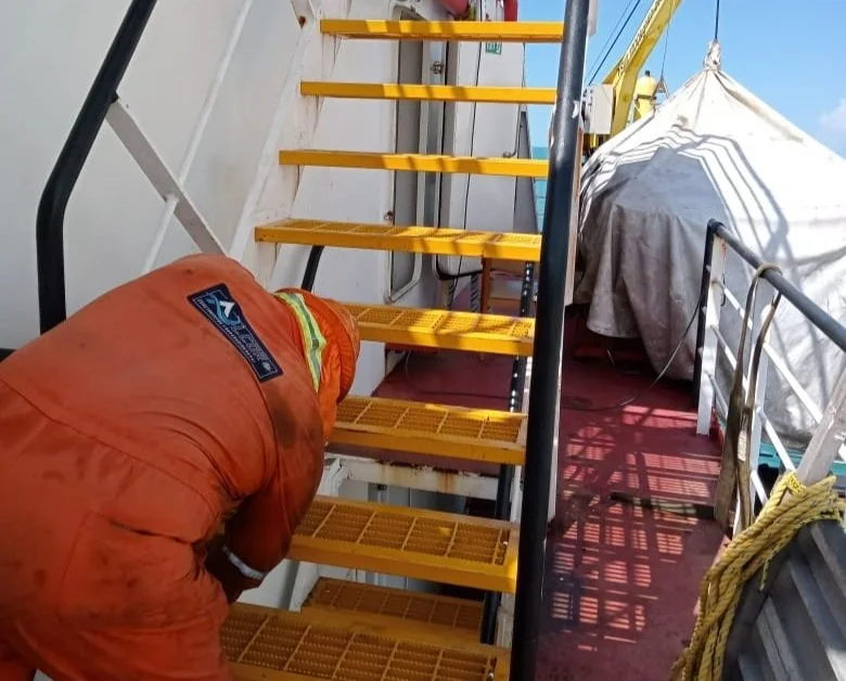 Persona en uniforme de trabajo inspeccionando o reparando una escalera amarilla en una estructura exterior, con equipo de protección y una tienda o cubierta blanca al fondo.