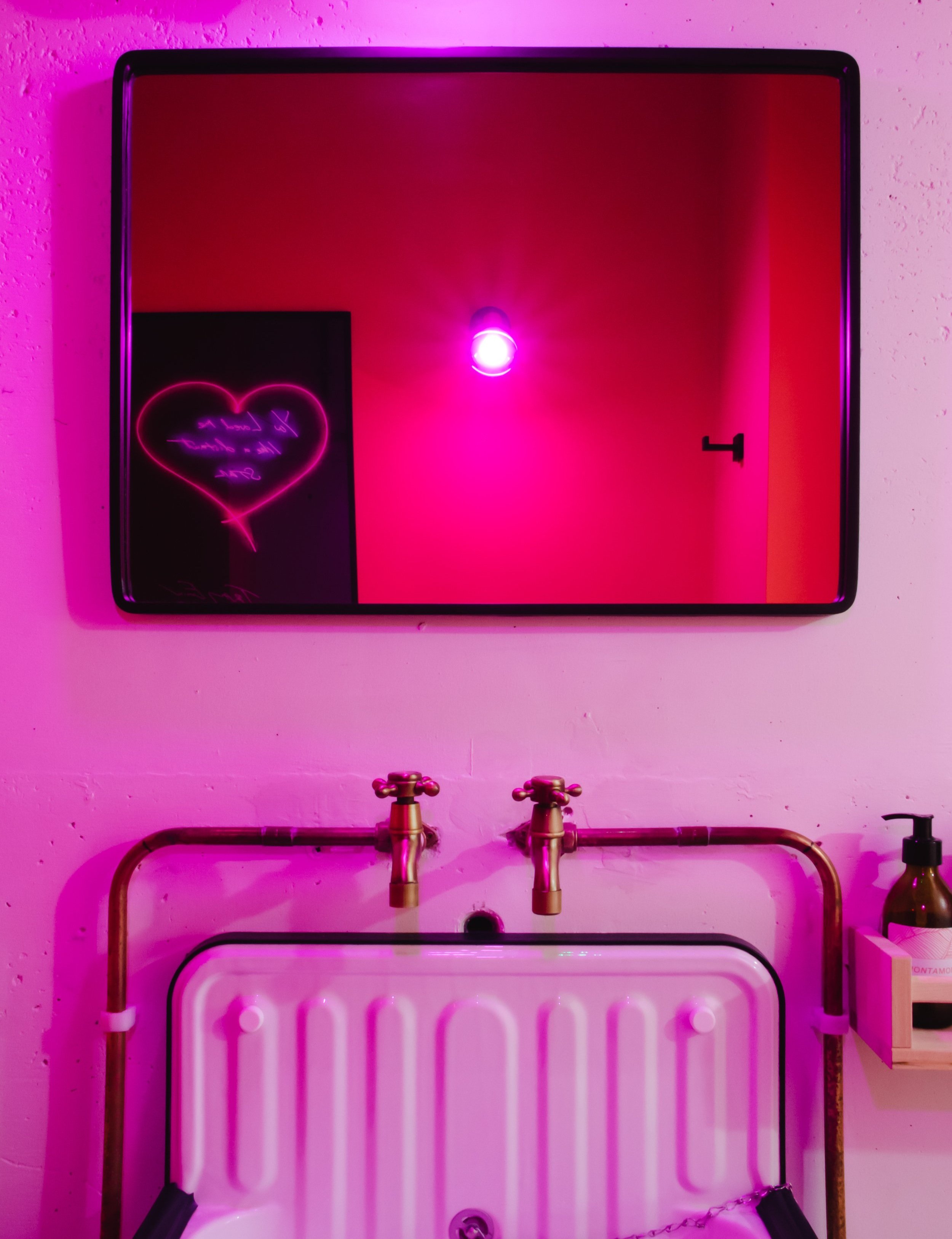 Bathroom with pink lighting and a mirror above a vintage basin with bronze faucets. The mirror reflects a pink-lit heart-shaped wall decoration and a light bulb.