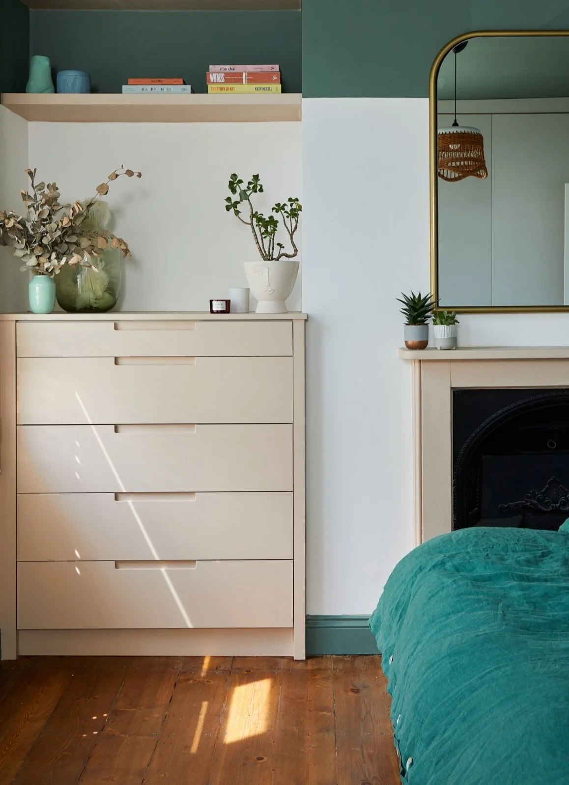 Minimalist bedroom with a beige dresser, decorated with plants and vases, books on the shelf, a large mirror, and a teal bedspread.
