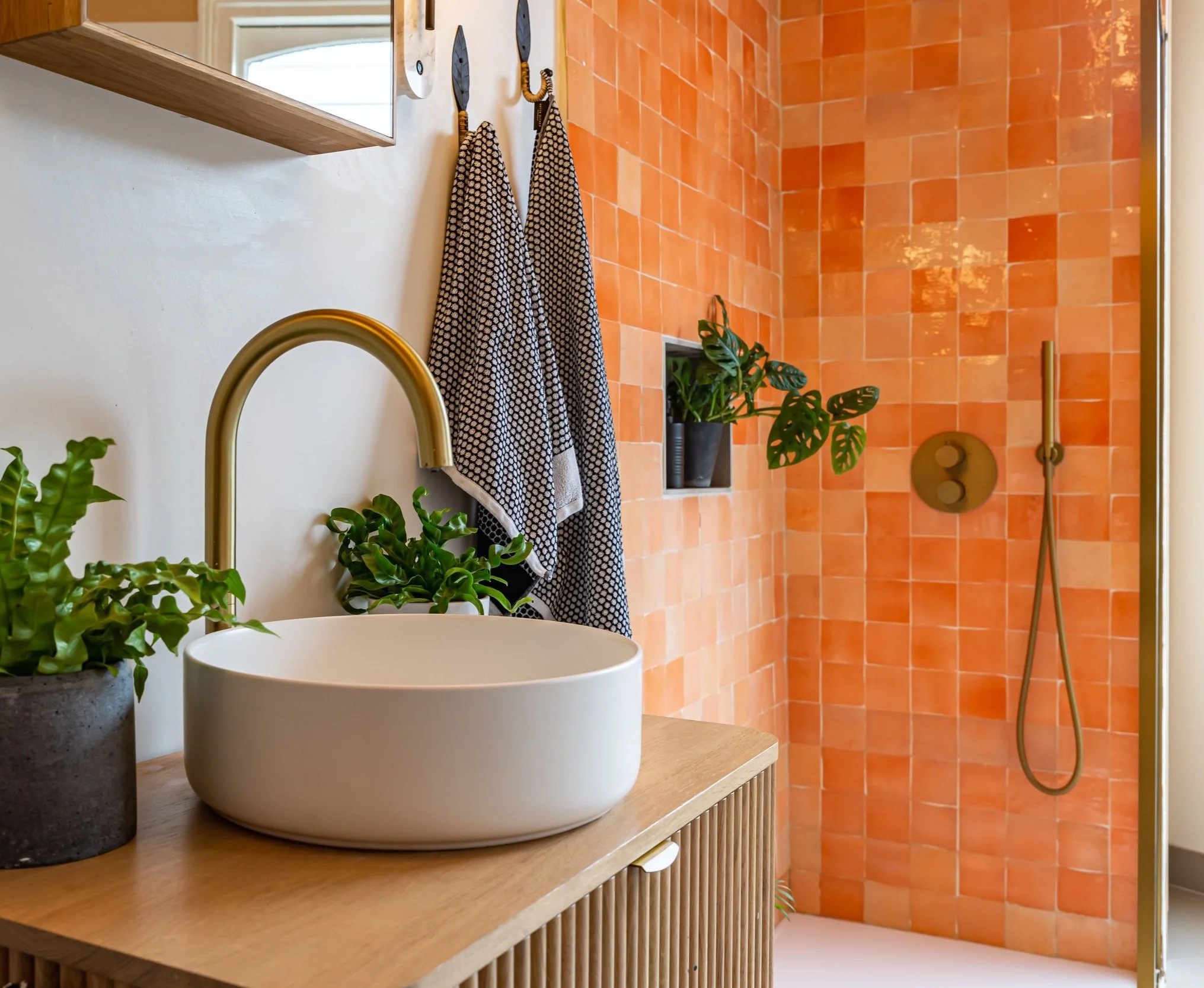 Modern bathroom with orange tiled shower, white vessel sink, gold faucet, green plants, and two towels hanging on hooks.