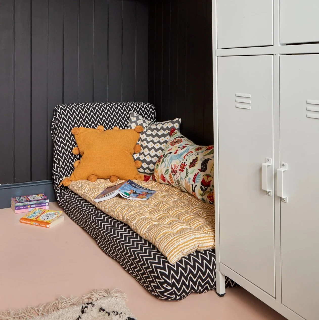 Cozy reading nook with zigzag-patterned floor cushion, colorful pillows, and a white storage cabinet. Books are spread on the floor and the cushion. Black wood panel walls add contrast.