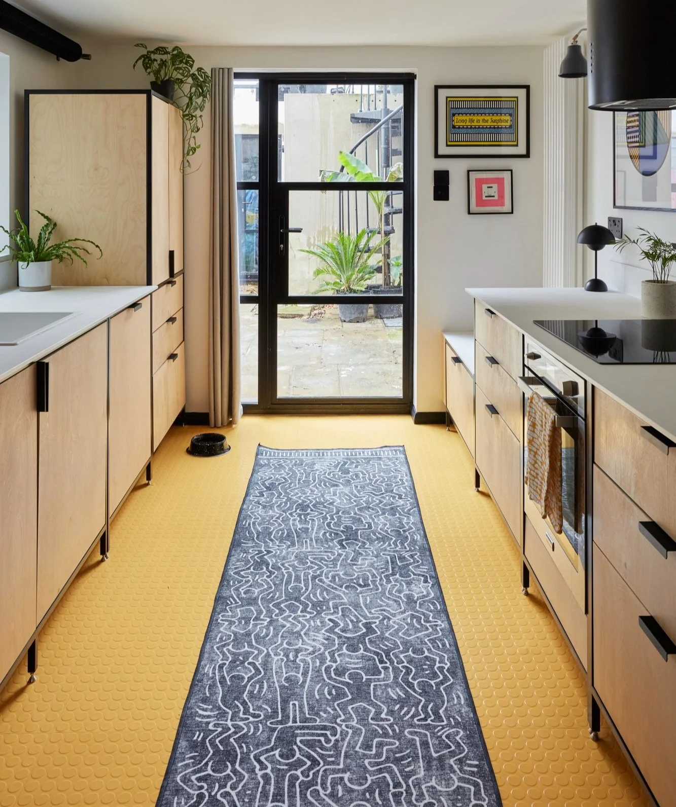 Modern kitchen with light wood cabinets, yellow hexagonal flooring, and a black and white patterned runner. Glass door to a patio with plants. Wall art and potted plants decorate the space.