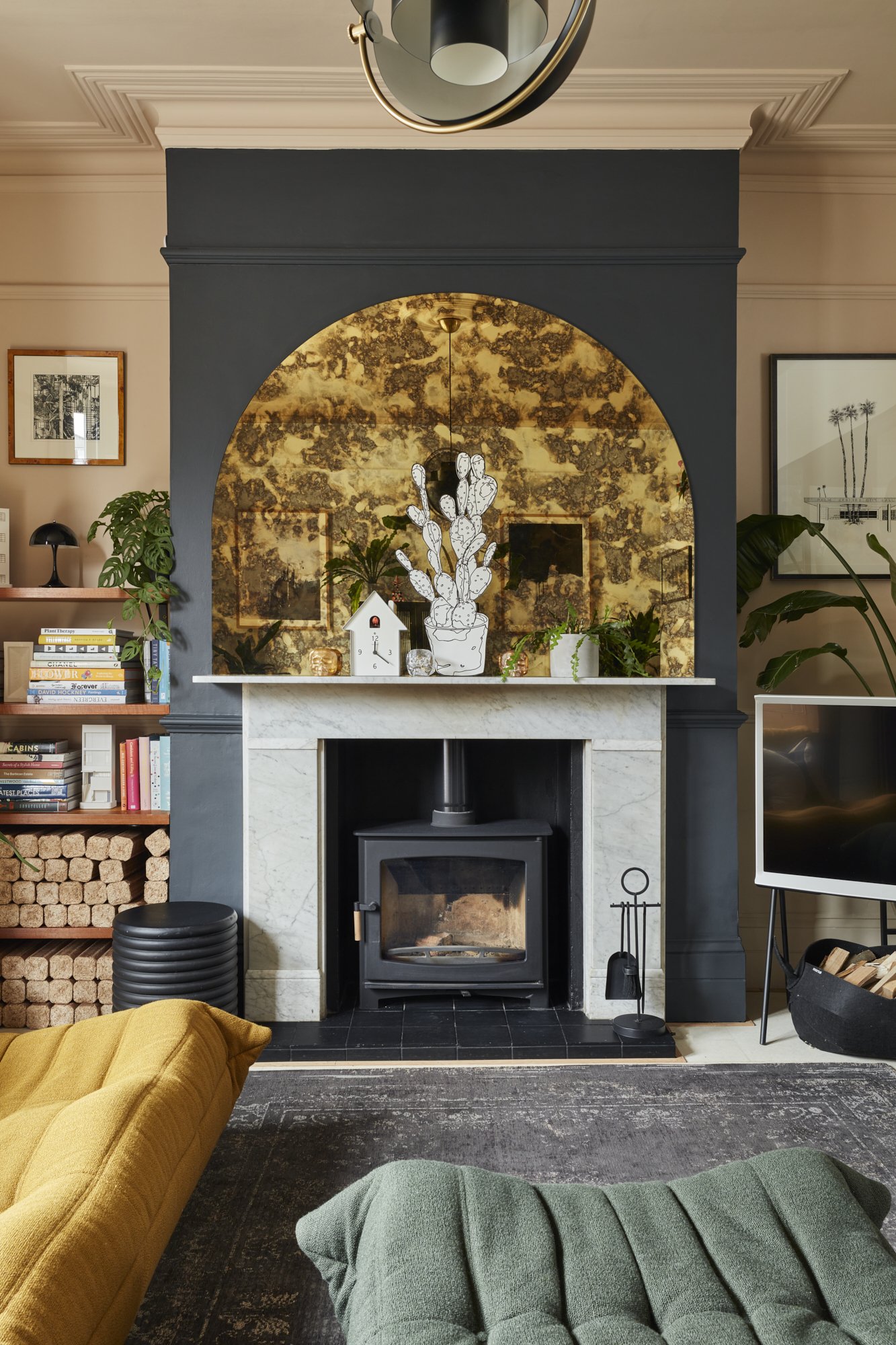 Living room with a black fireplace, marble mantle, decorative mirror, and wood-burning stove. Bookshelves with books and decor items flank the fireplace. Yellow and green cushions on the floor. A black stool and firewood storage are nearby.