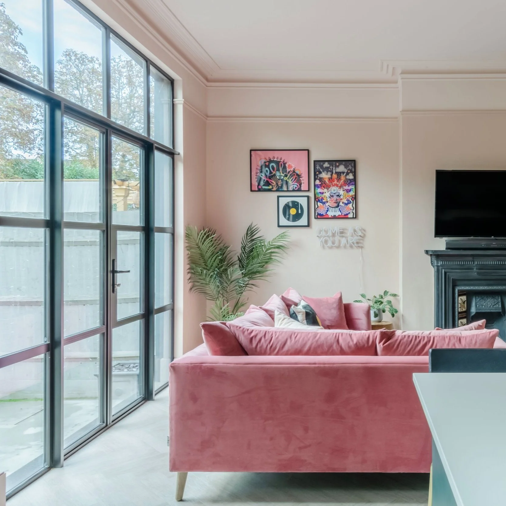 Modern living room with a pink velvet sofa, decorative wall art, and a black TV above a fireplace. Large glass doors on the left, plants, and minimalistic decor.