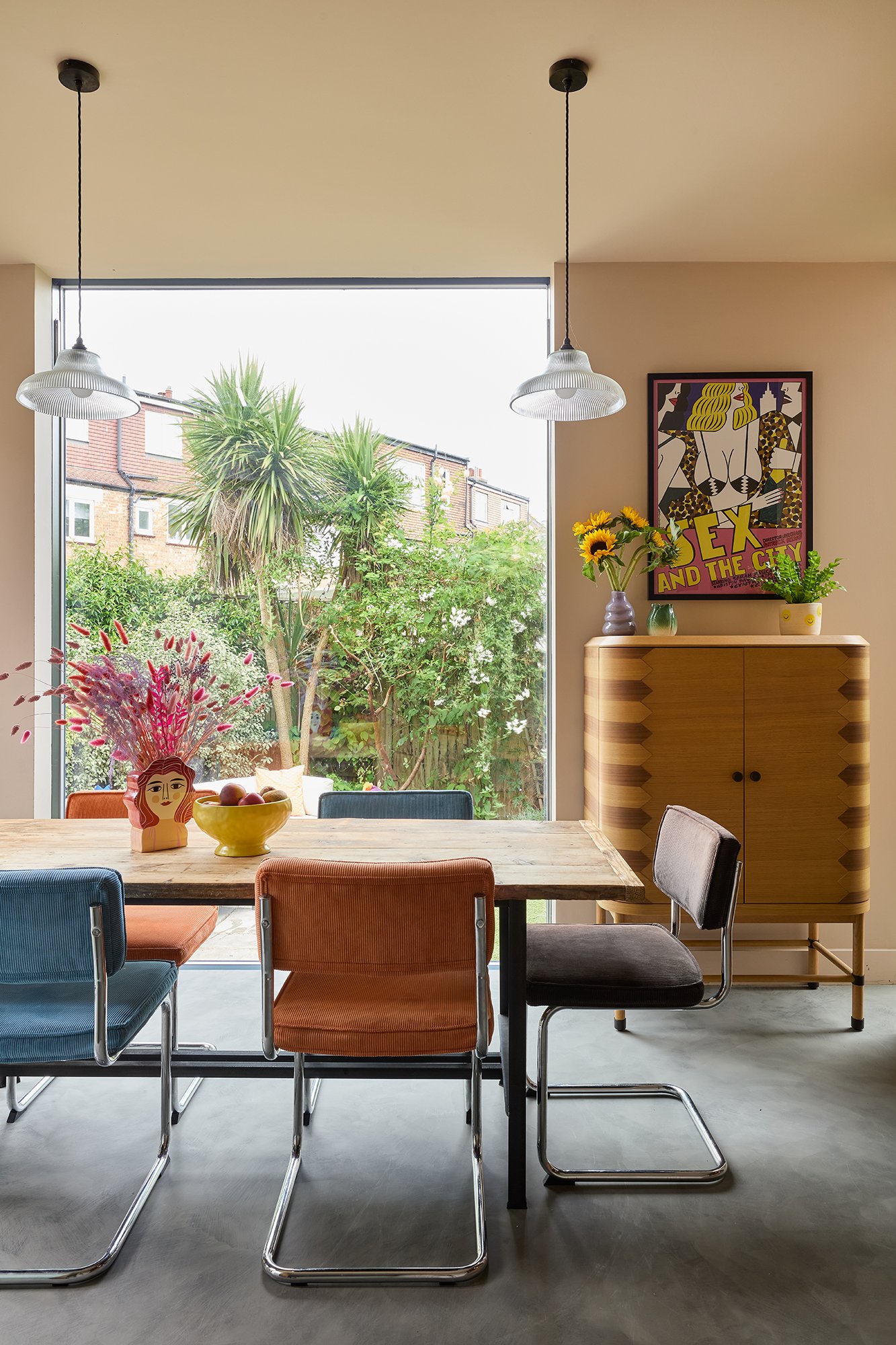 Modern dining room in Haringey with a wooden table, colourful chairs, and large window overlooking a garden. Two pendant lights hang above the table. A poster and plants are on a wooden cabinet.