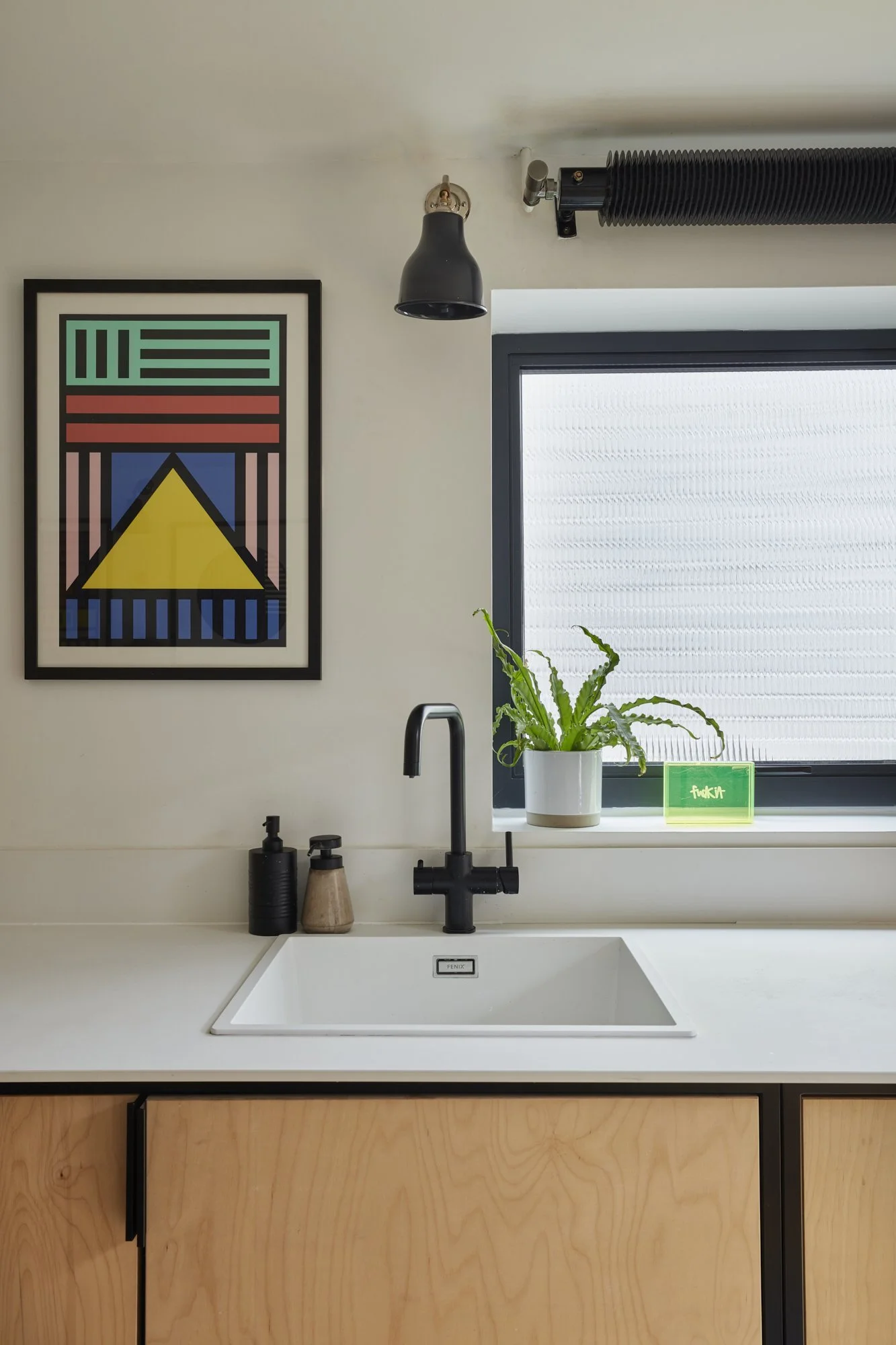 Modern kitchen sink area with a white countertop, black faucet, and wooden cabinets. A potted plant is on the window sill, next to a green box. A black wall lamp and abstract framed art are above the sink.