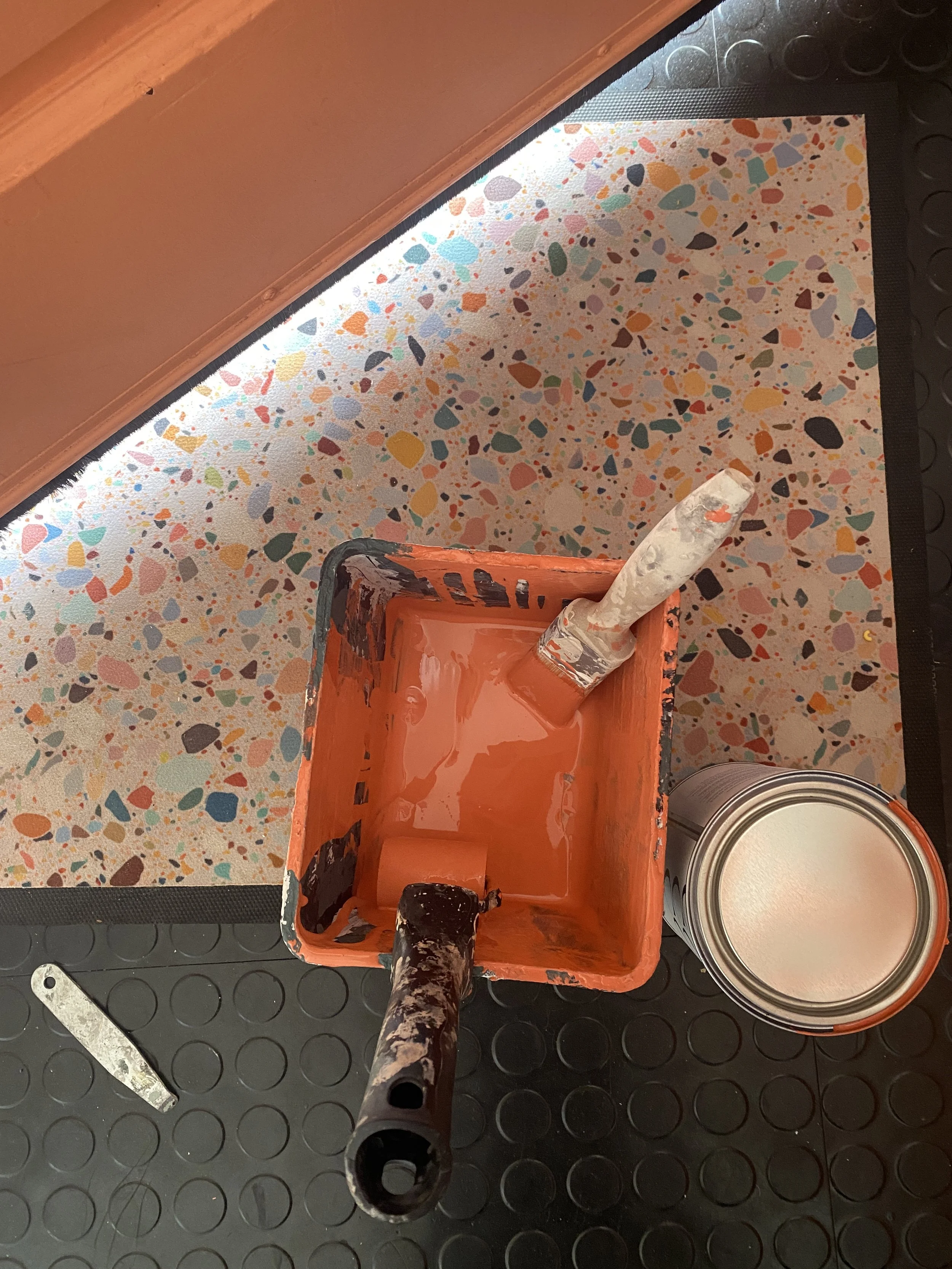 Paint tray with orange paint, two paintbrushes, an open paint can, on a colorful terrazzo-patterned surface.