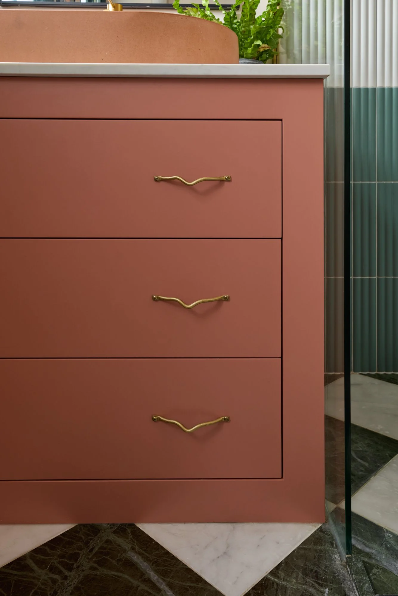 Modern bathroom vanity with pink drawers, brass handles, and a white countertop. Visible textured backsplash and partial greenery decor.