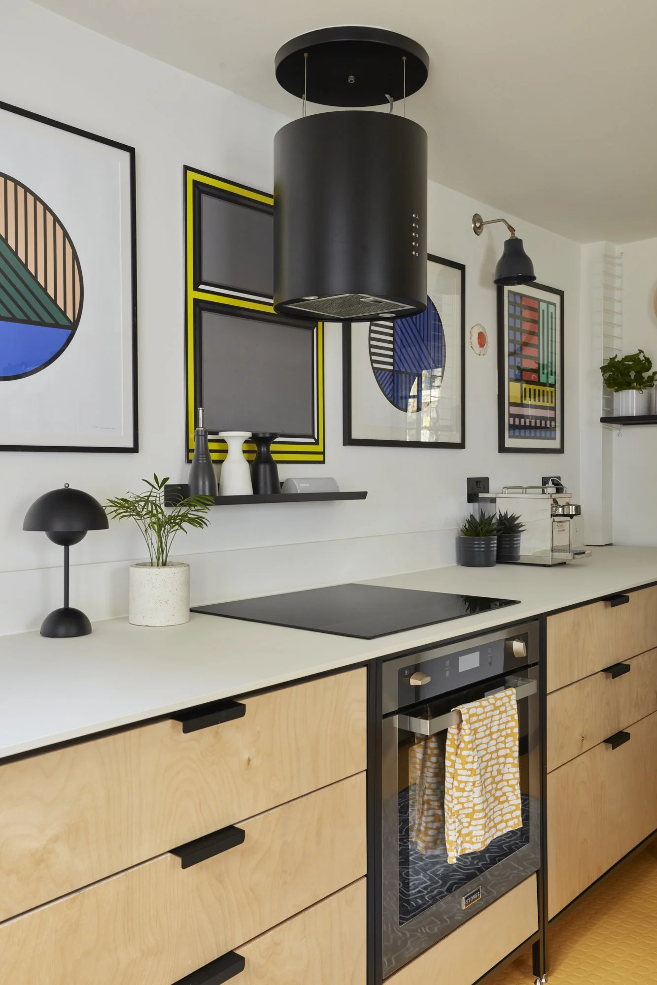 Modern kitchen interior with minimalist design, featuring light wood cabinets, black handles, a black overhead range hood, and a stovetop oven. Wall decor includes framed geometric art prints. The countertop displays a small black lamp, a potted plant, and decor items.