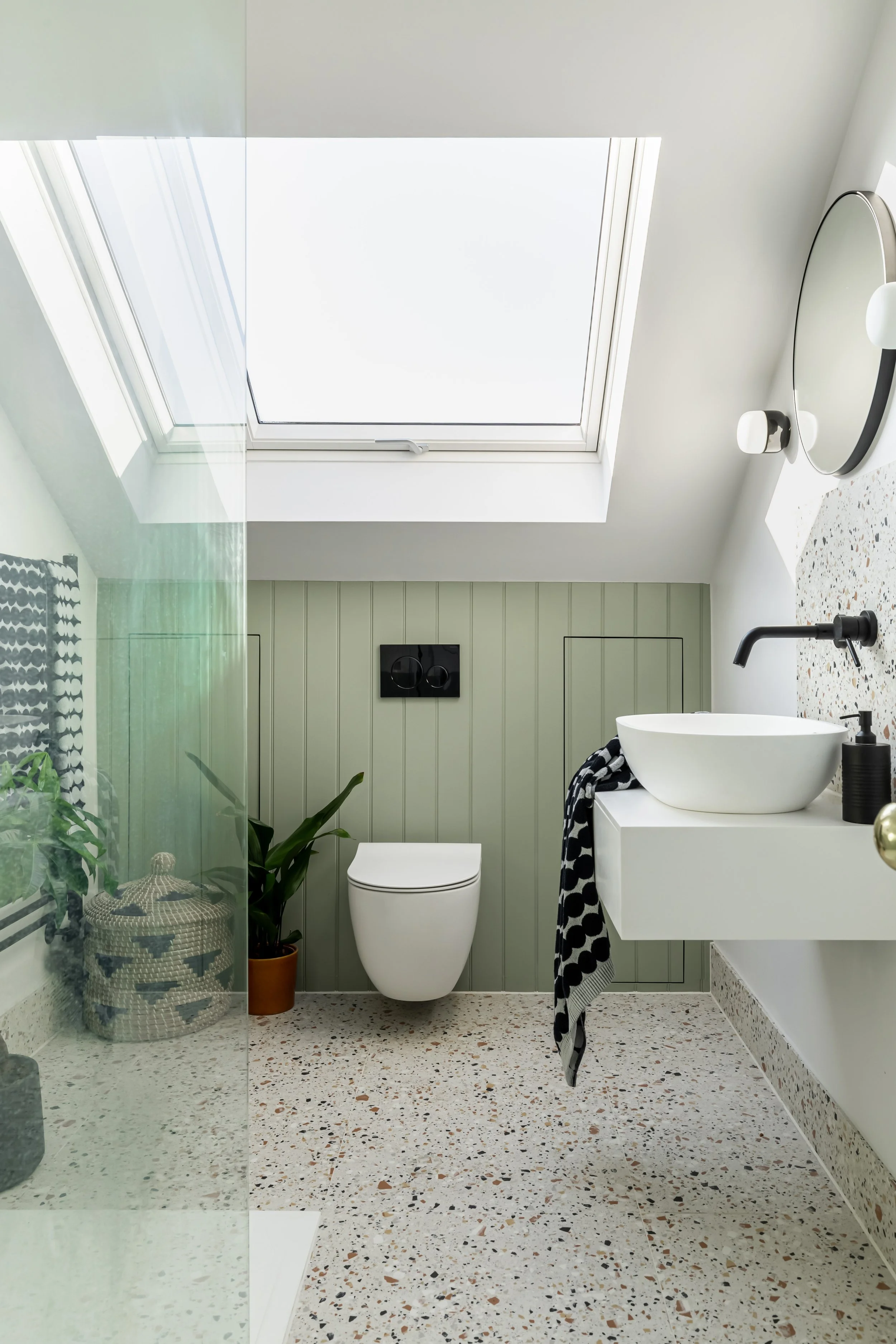 Modern bathroom with terrazzo flooring, wall-mounted toilet, round vessel sink, black faucet, skylight, and plants.