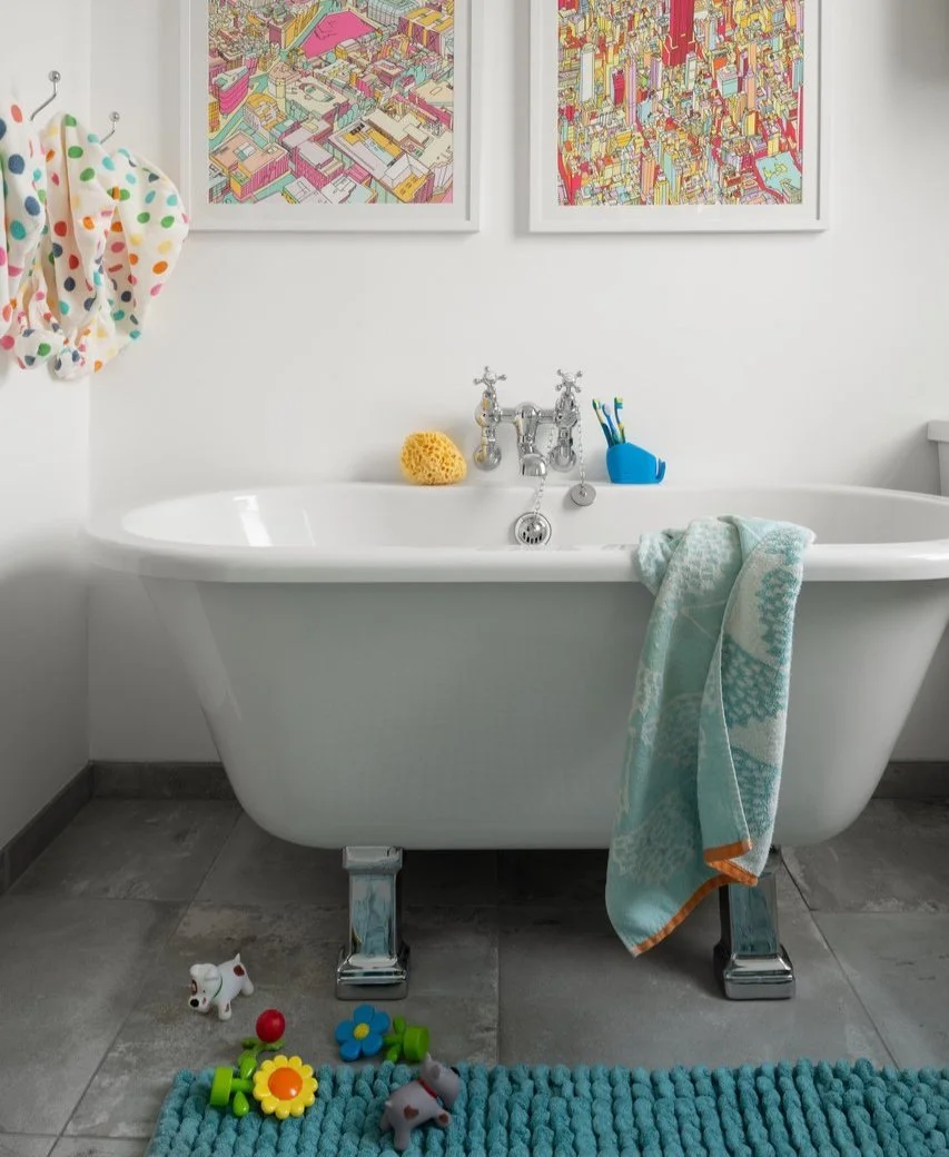 Bathroom with a freestanding white bathtub, colorful children's toys on the floor, blue textured bath mat, blue towel hanging on the tub, two vibrant cityscape artworks on the wall, and a wall hook with a polka dot bathrobe.