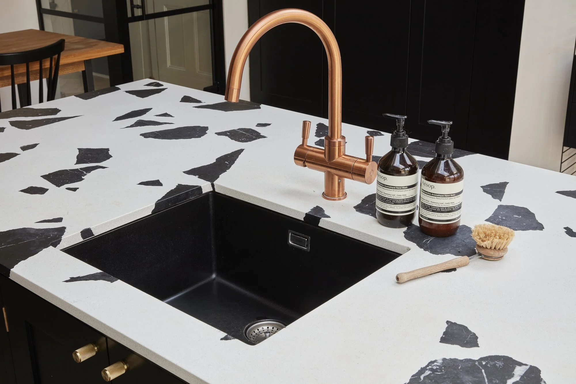 Kitchen countertop with black and white terrazzo pattern, copper faucet, black sink, hand soap bottles, and a wooden scrub brush.