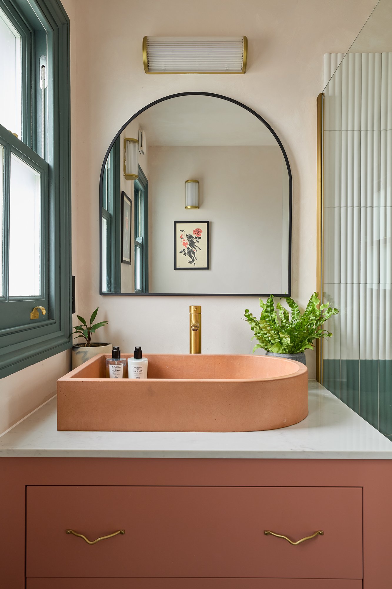 Modern bathroom with pink sink and gold faucet, large mirror, potted plants, wall art, and wall light.