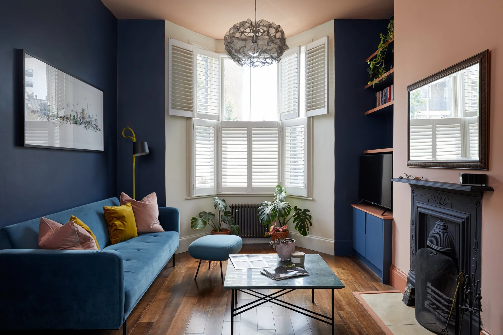 Modern living room with blue sofa and yellow pillows, wooden floor, large window with shutters, black fireplace, and glass coffee table with magazines.