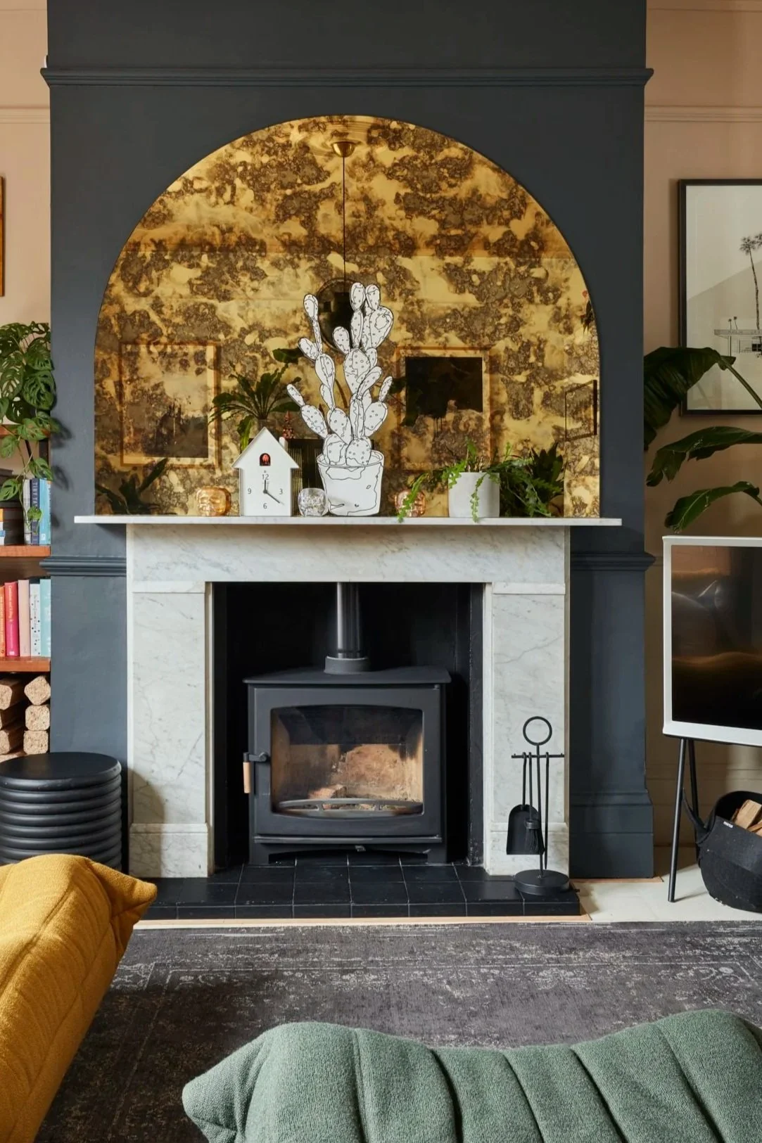 Living room fireplace with black wood stove, marble surround, dark framing, and decorative items including a cactus drawing, clock, and plants on the mantle. There are armchairs, a television, and shelves with books nearby.
