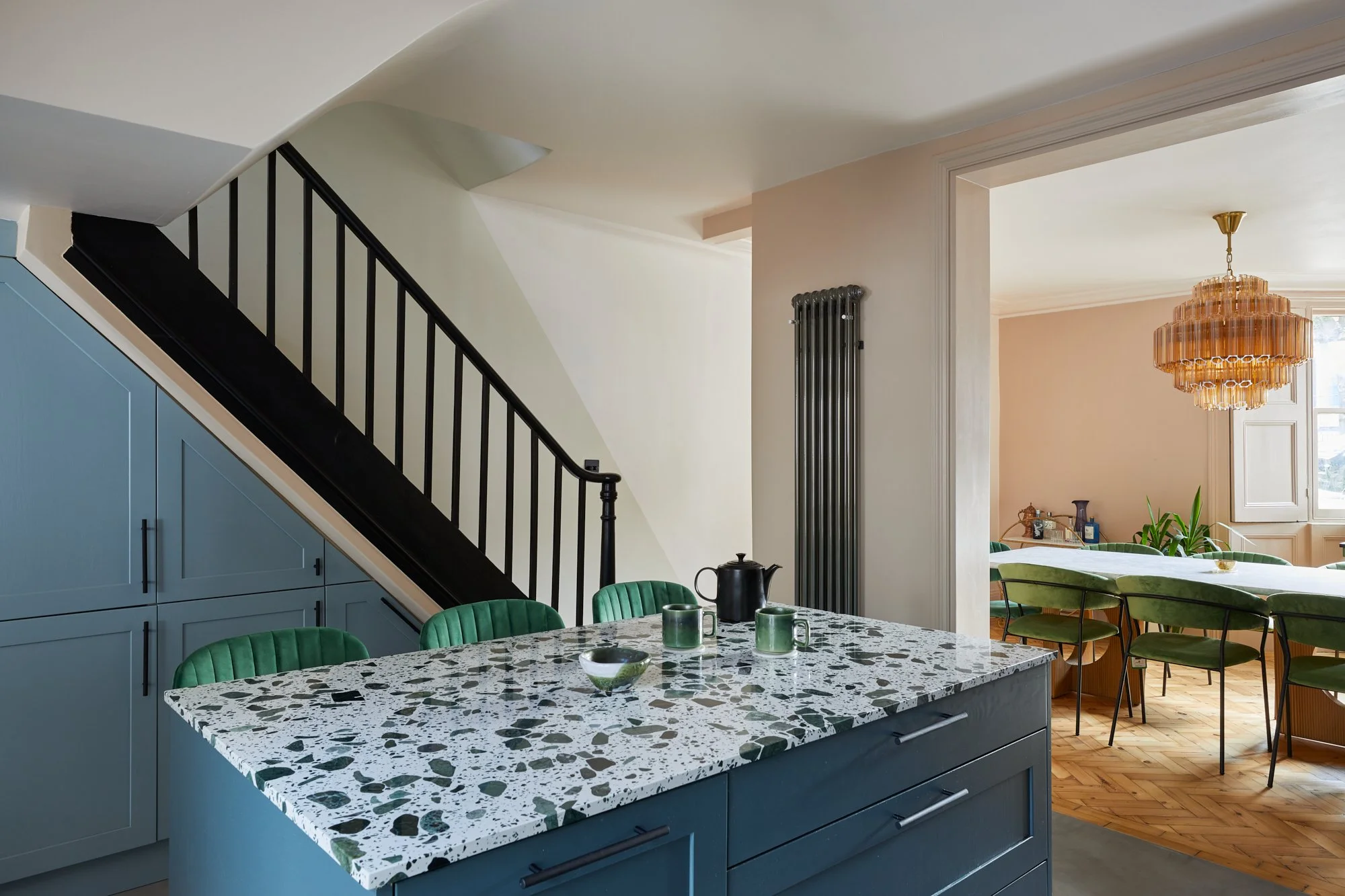 Modern kitchen interior with terrazzo countertop, blue cabinetry, green velvet chairs, black staircase railing, and a glimpse into a dining area with a pendant chandelier.