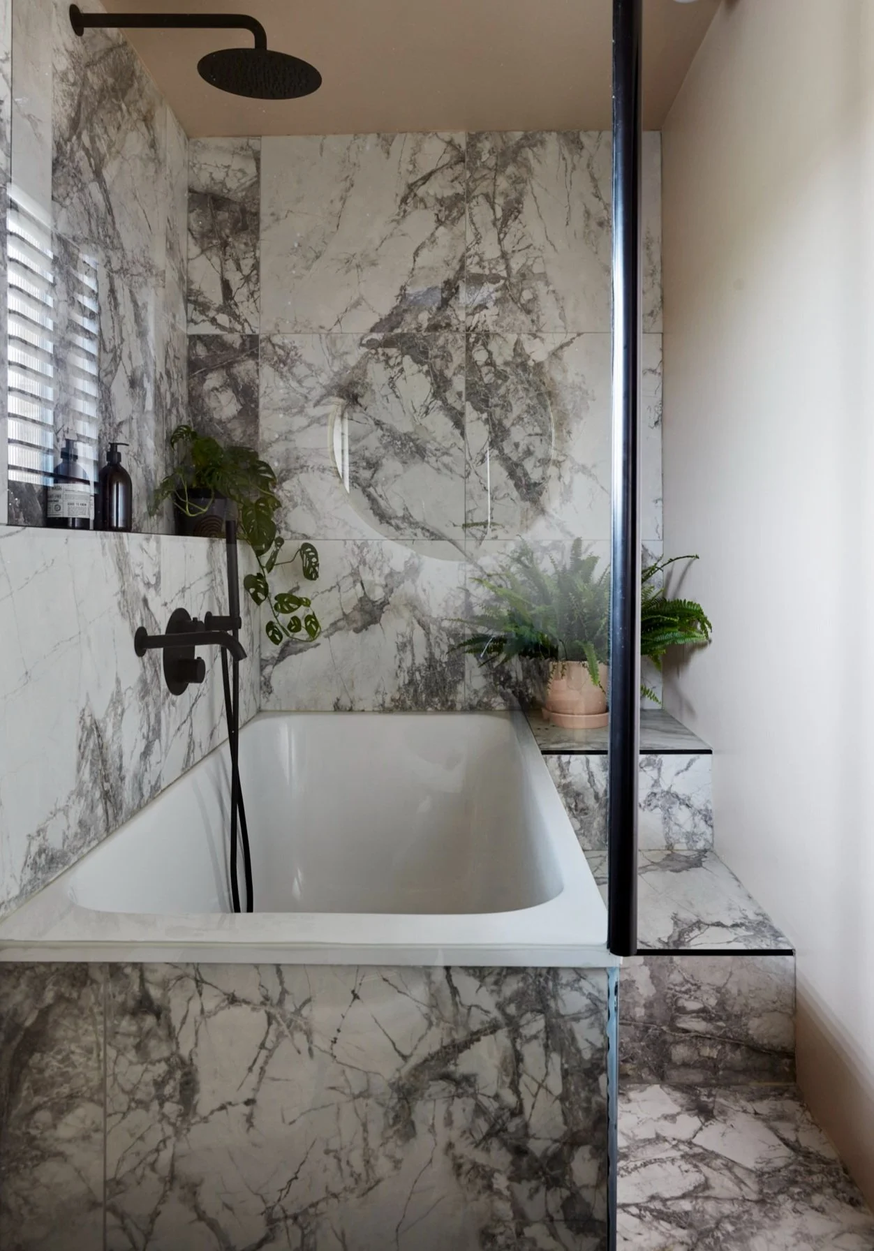 Marble bathroom with a white bathtub, black faucet, and rainfall showerhead. The walls and steps are covered in gray marble. Potted plants decorate the area.
