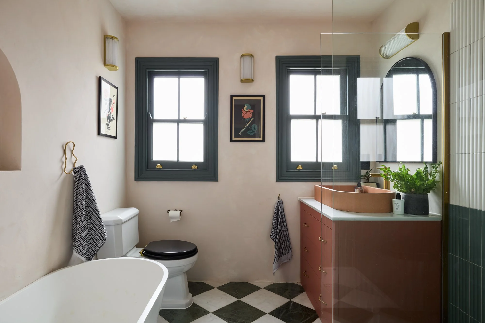 Modern bathroom with a black and white checkered floor, a freestanding bathtub, a toilet with a black seat, green-framed windows, a red vanity with a round sink, and a mirror. A potted plant and decorative artwork are present.