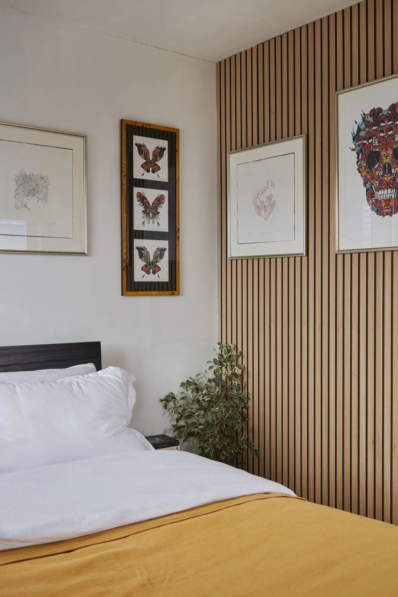 Bedroom corner with wood paneling and wall art, including butterfly and skull prints, next to a bed with white bedding and a yellow blanket.