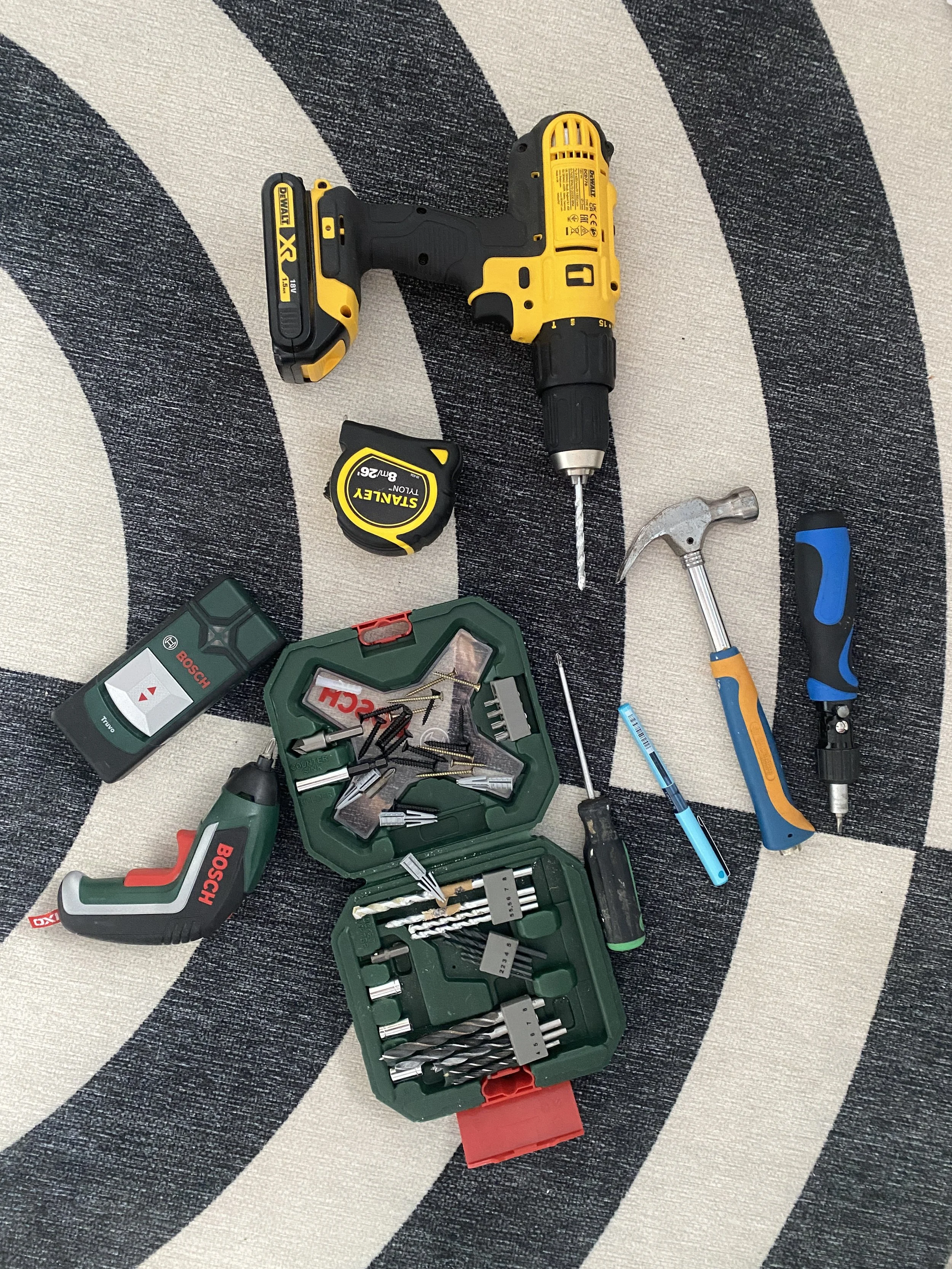 Assorted tools on a patterned rug, including a yellow cordless drill, tape measure, hammer, screwdriver, utility knife, drill bits in a case, and small power tools.