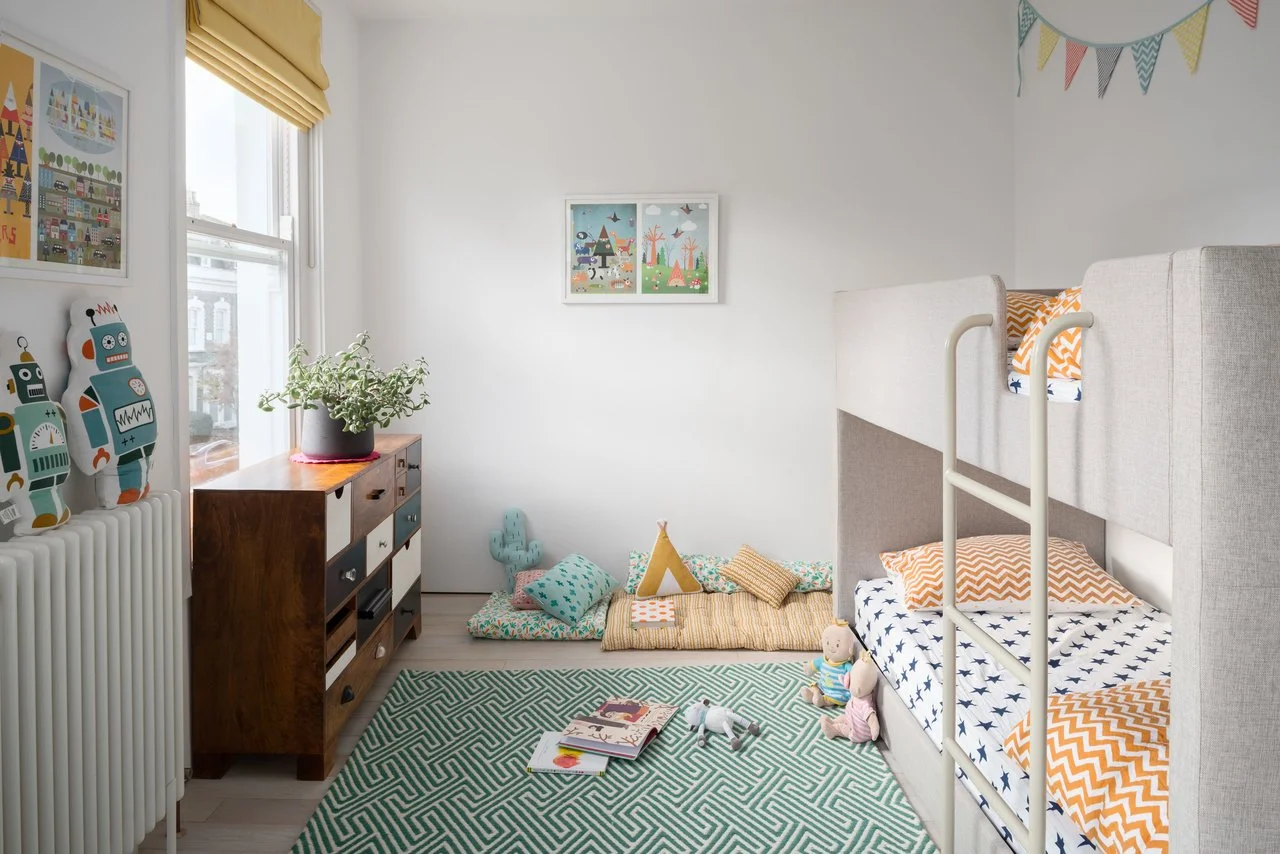 Kids' room with bunk bed, colorful bed linens, wooden dresser, toys, books on a geometric rug, and wall art.
