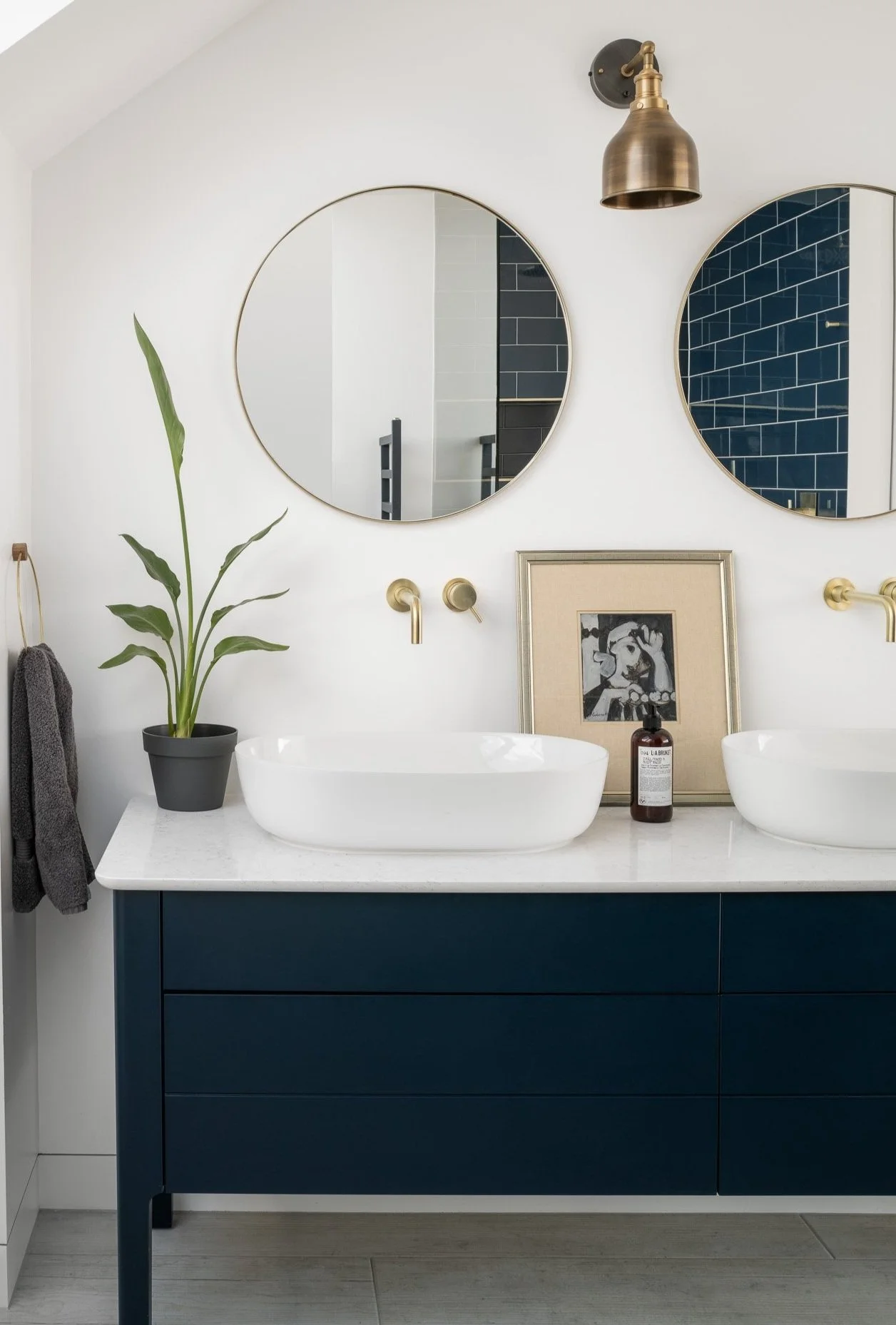 Modern bathroom with two round mirrors, white vessel sinks, gold faucets, a small plant, and framed artwork on a blue vanity.