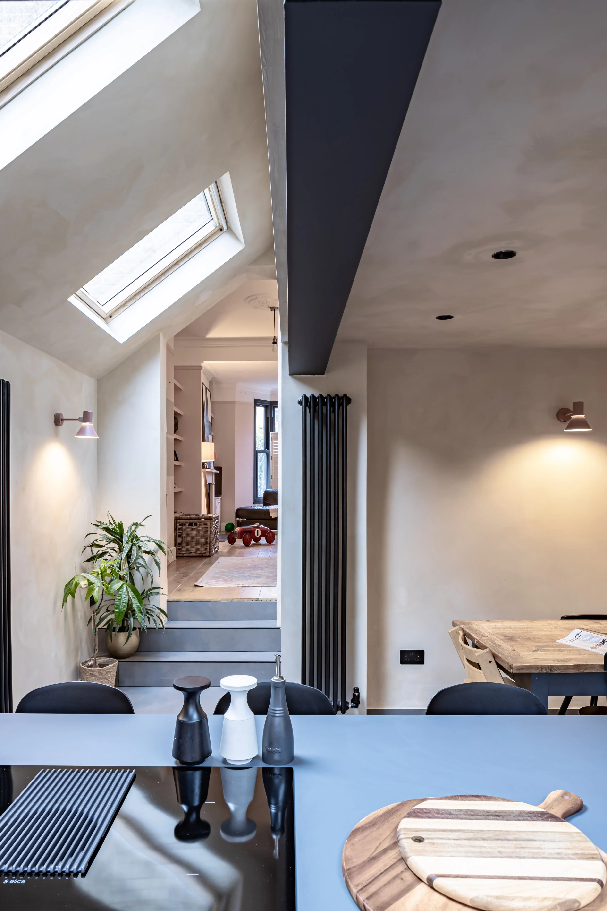 A modern kitchen and dining area with a skylight, light-colored walls, and a wooden dining table. There are three containers on a sleek countertop, a cutting board, and a potted plant near the steps leading to a cozy living space. Minimalist decor includes wall-mounted lights and vertical radiators.