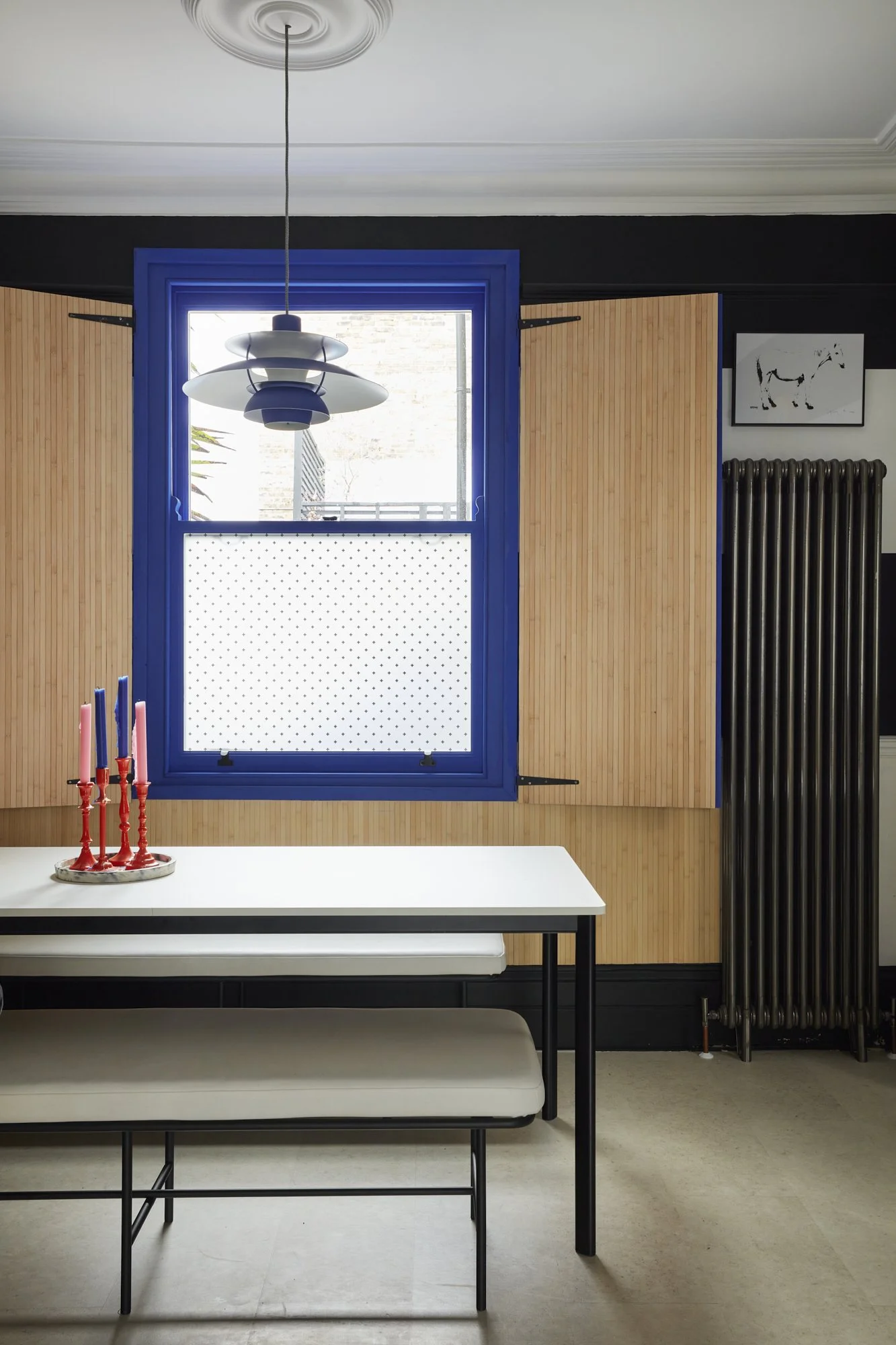 Modern dining room with a white table and bench seating, red and pink candles on the table, bamboo paneling, a blue window frame, a pendant light, and a black radiator.