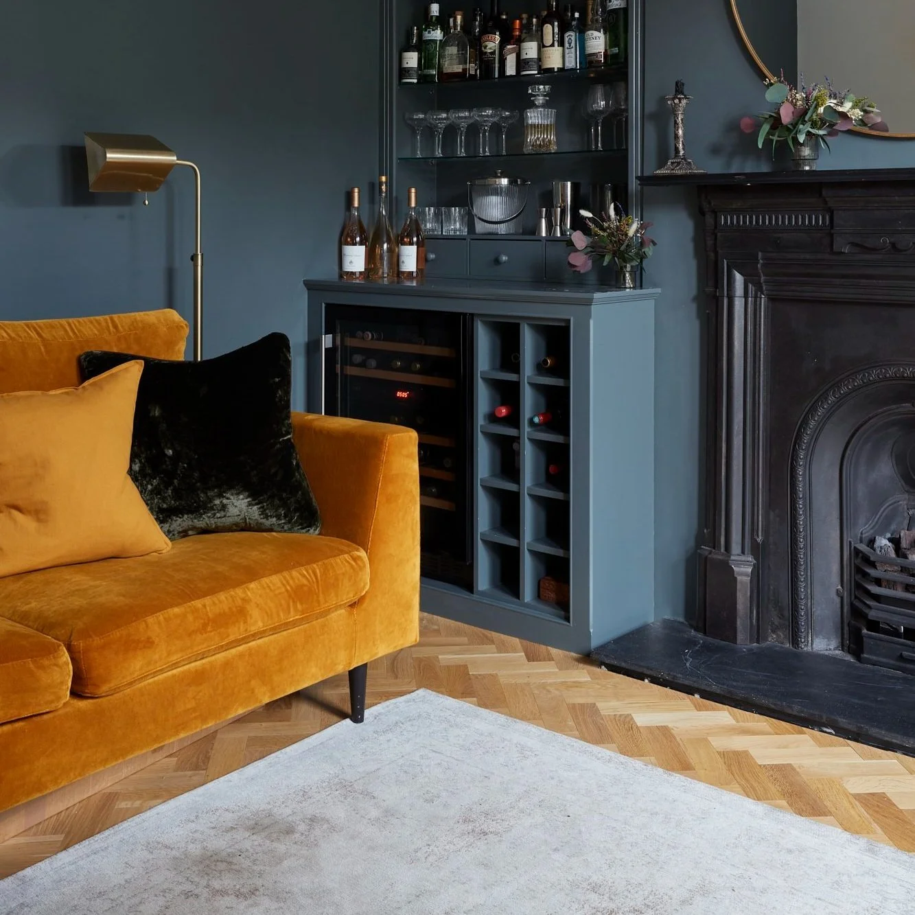 Cozy living room with a mustard yellow sofa, dark blue walls, a bar cabinet with wine storage, and a black ornate fireplace.
