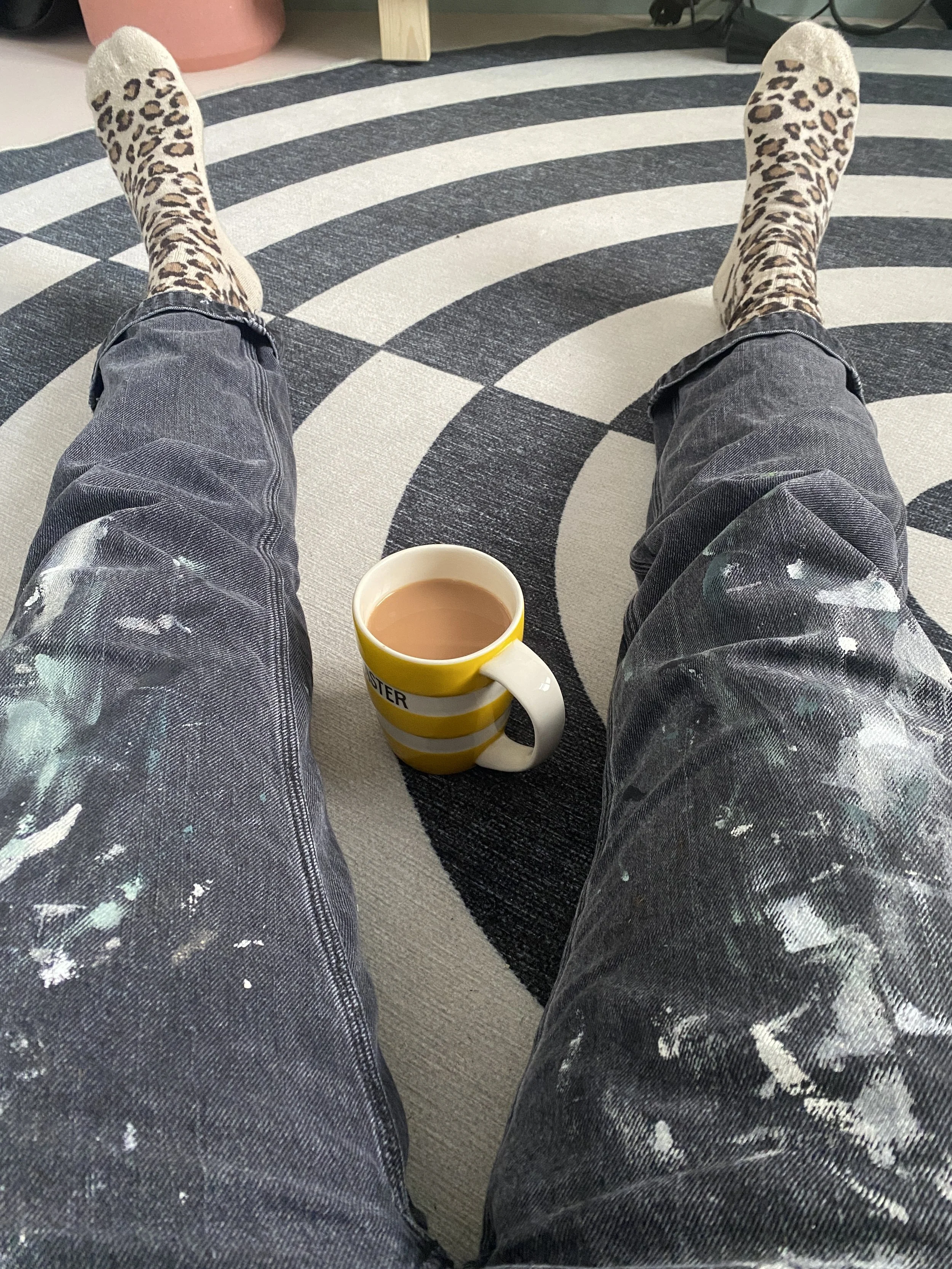 Person wearing leopard print socks and paint-splattered jeans sitting on a round black and white striped rug with a yellow striped mug of coffee between their legs.