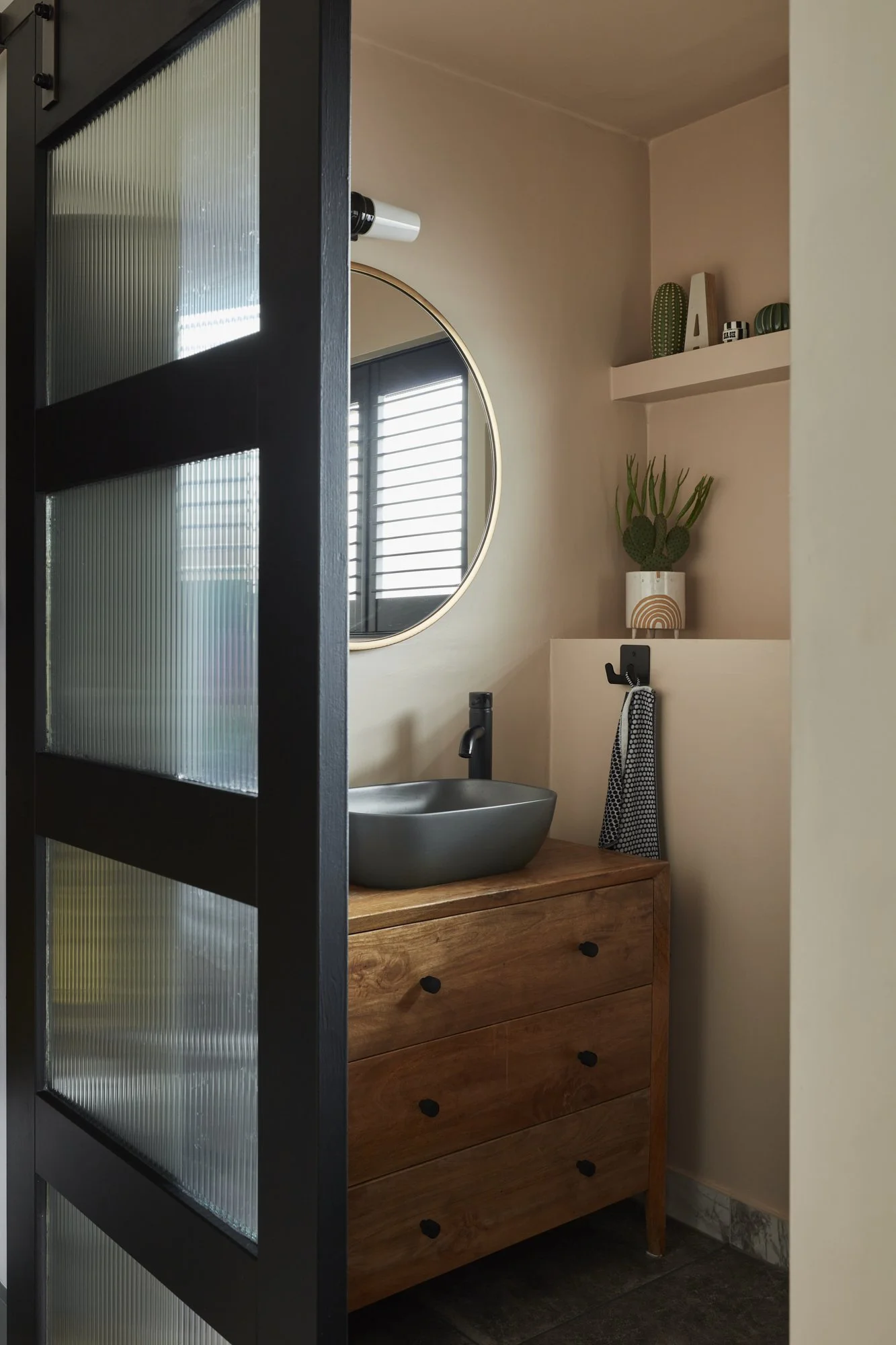 Modern bathroom with wooden vanity, vessel sink, round mirror, sliding glass door, and decorative cactus.