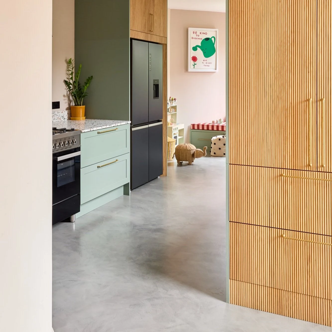 Modern kitchen with mint green cabinets, black refrigerator, wood paneling, terrazzo countertop, and indoor plants. Adjacent playroom visible with toys and a wall poster.