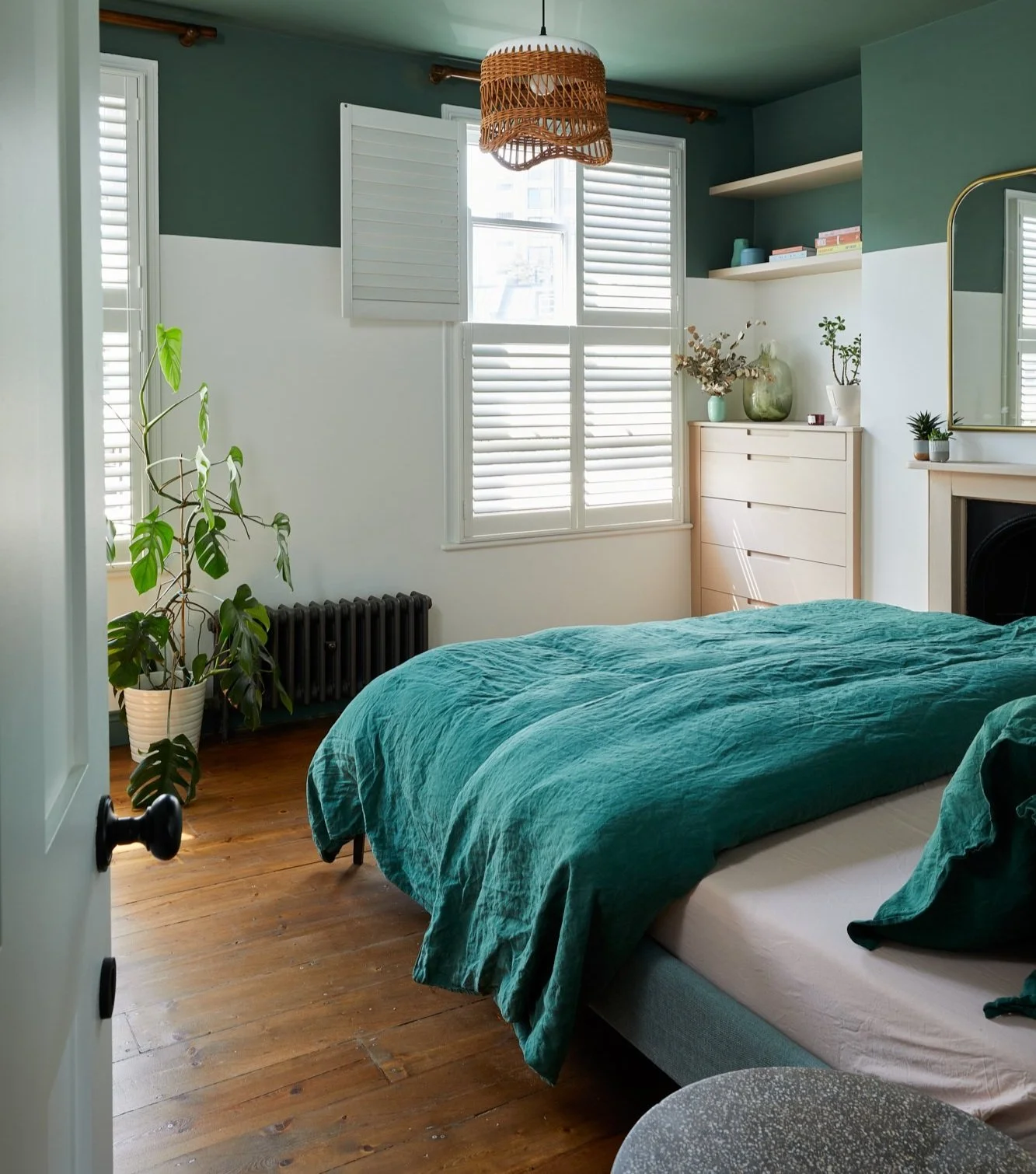 Bedroom with green and white walls, wooden floor, bed with green bedding, wicker light fixture, potted plant, chest of drawers, and window shutters.