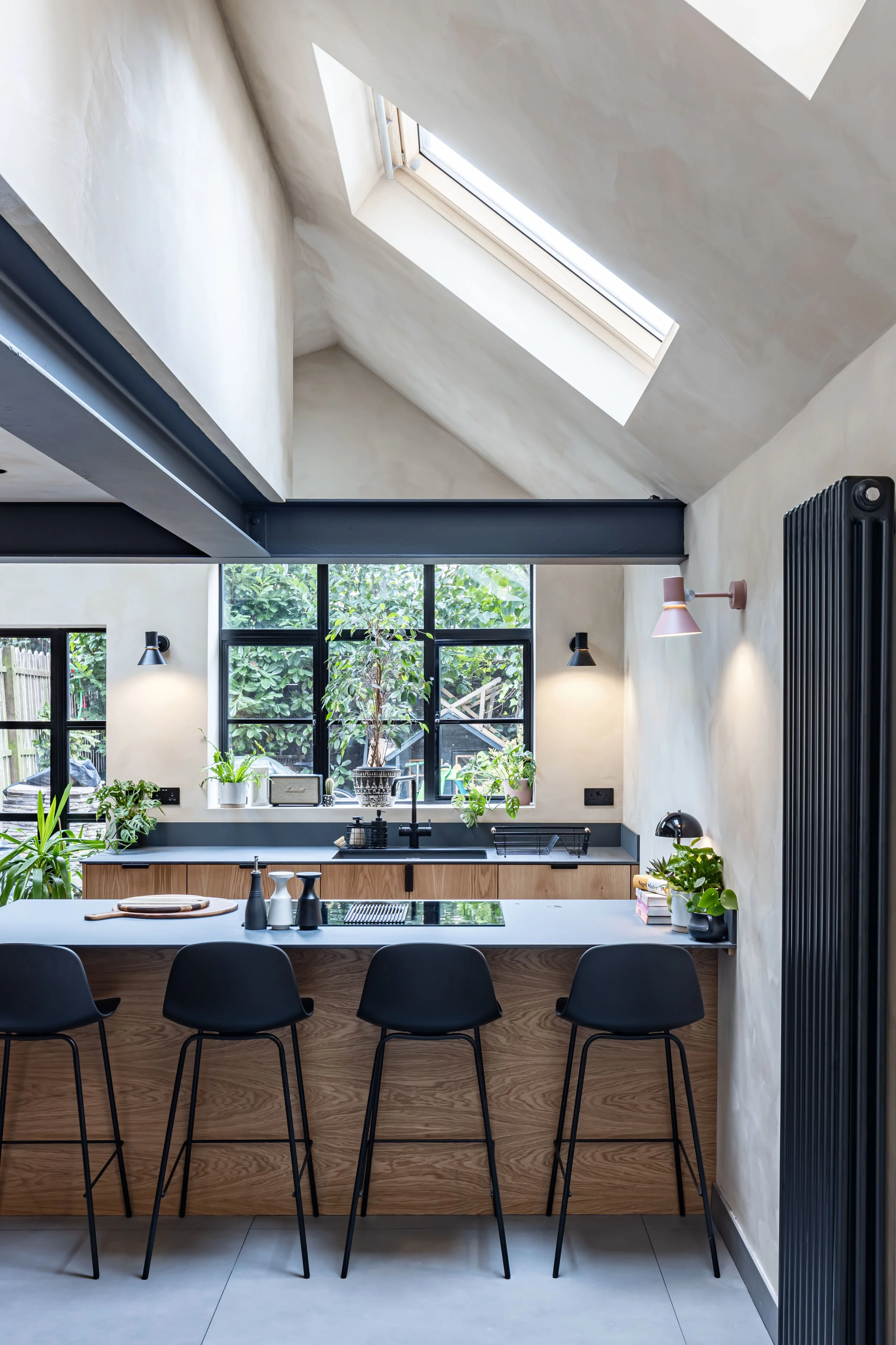 Modern kitchen interior with skylight, black bar stools at a wooden island, plants, and large windows looking out to greenery.
