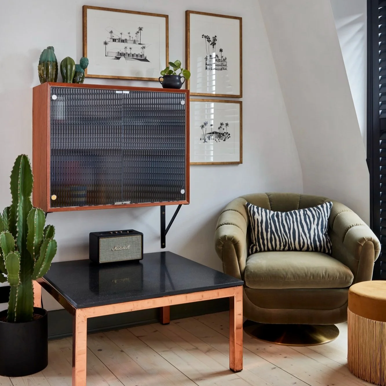 Modern living room corner with a green armchair, zebra print pillow, wall-mounted cabinet, black table, and plants. Art frames with black and white illustrations on the wall.