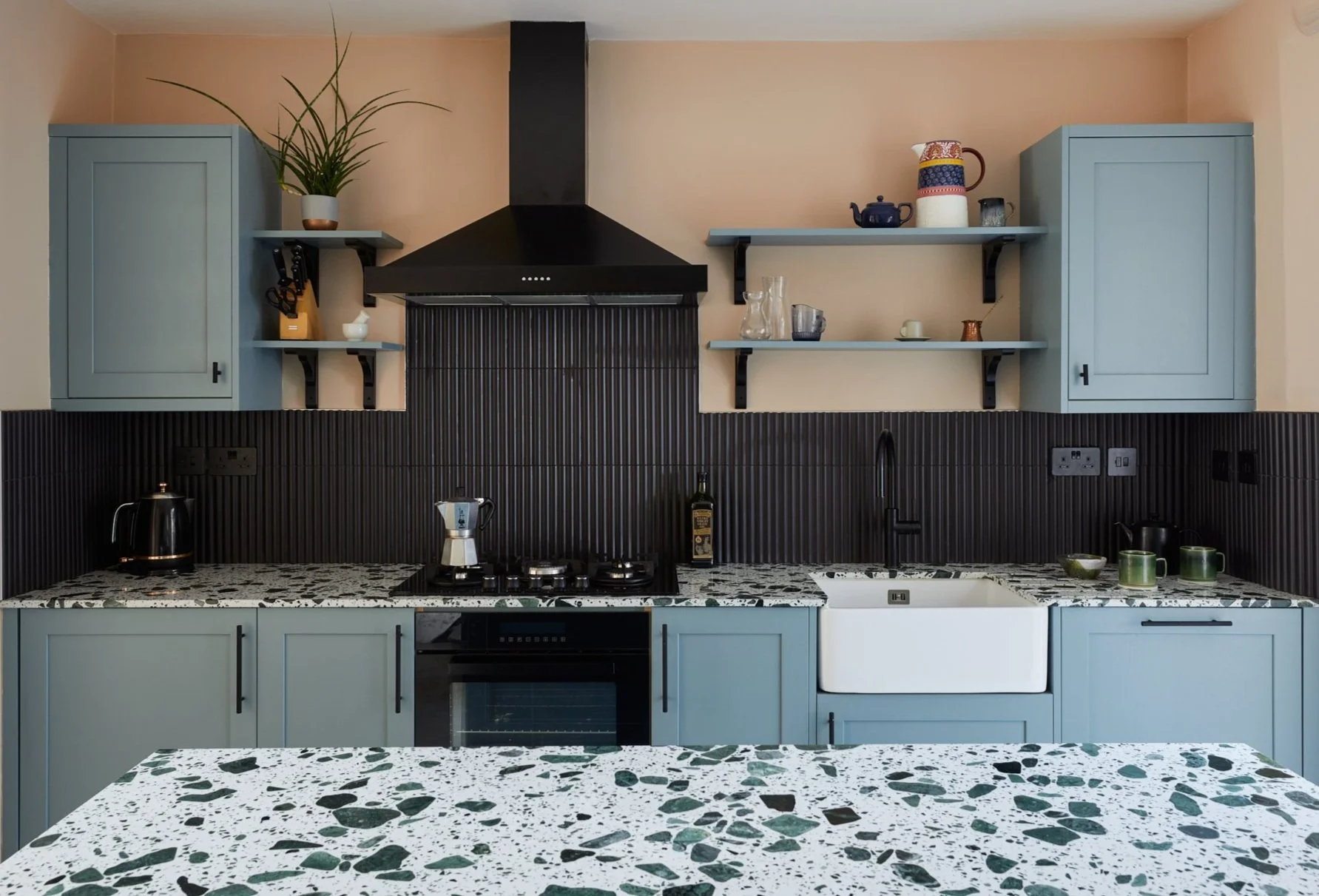 Modern kitchen with blue cabinets, black range hood, terrazzo countertops, farmhouse sink, and shelves with various kitchen items.