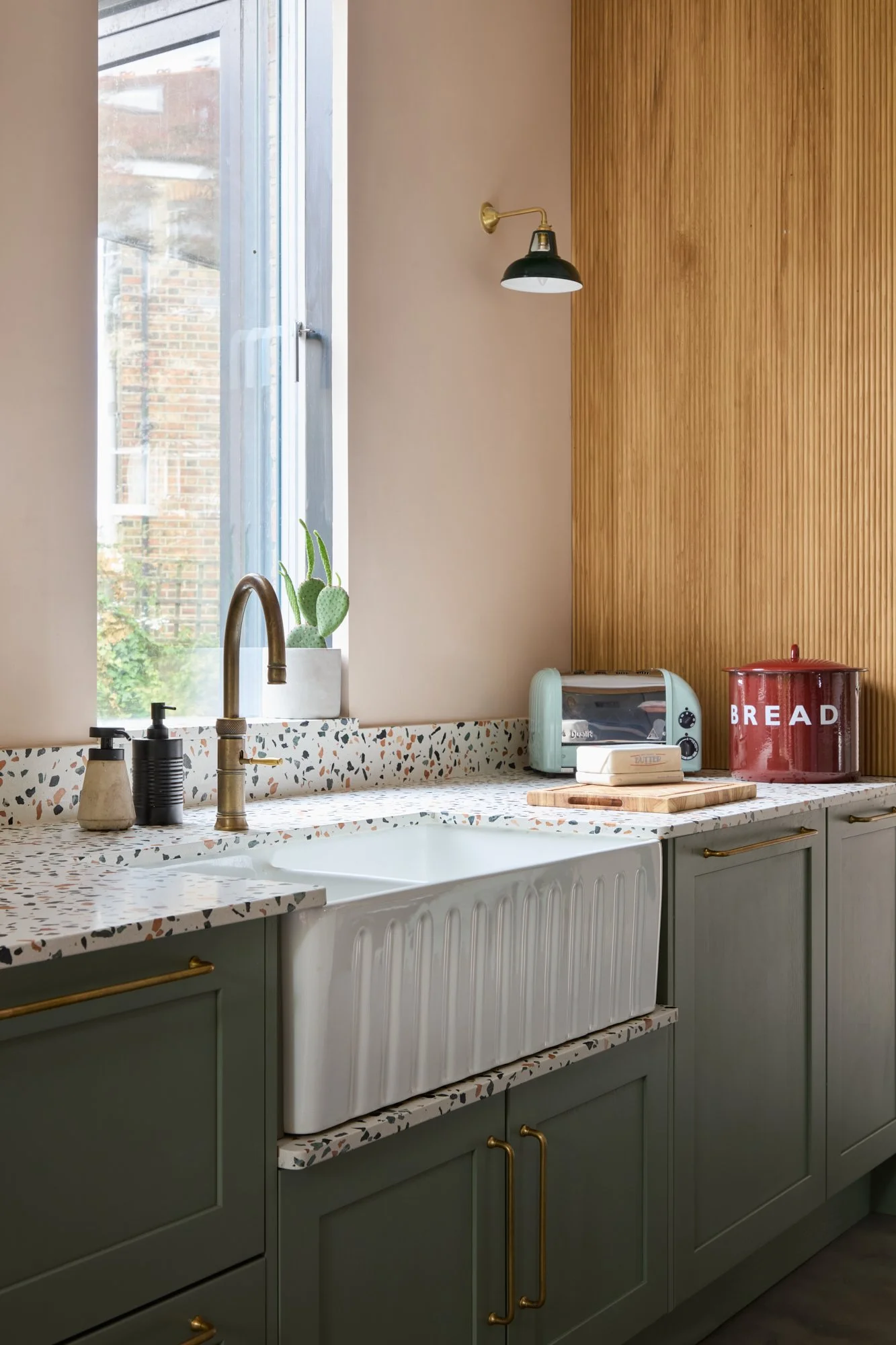 Modern kitchen with green cabinets, terrazzo countertops, and a farmhouse sink. Brass faucet with soap dispensers nearby and a potted cactus by the window. Red bread box and toaster on the counter. Wooden accent wall and wall-mounted light fixture above.