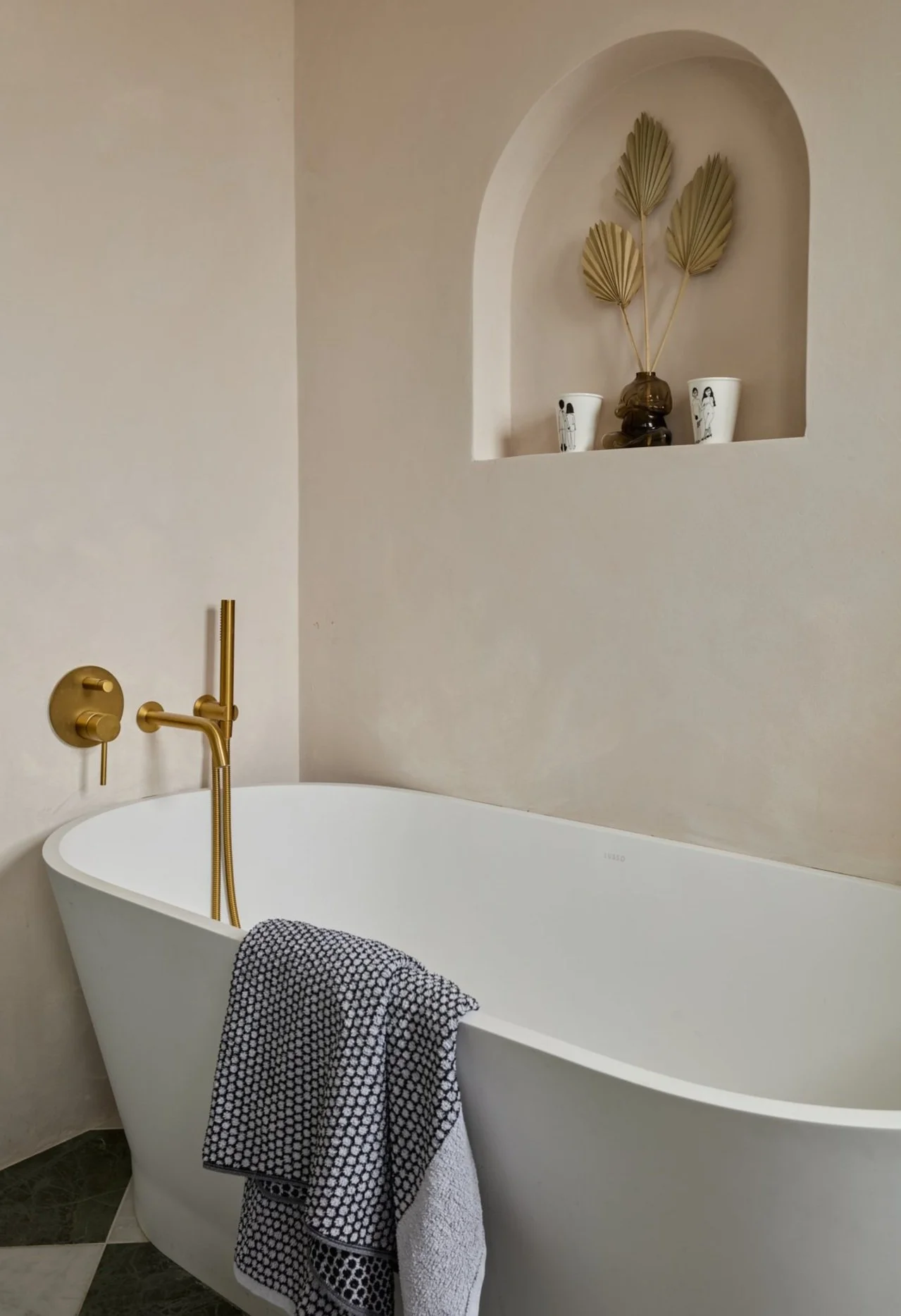 Minimalist bathroom with a white freestanding bathtub, gold faucet, and a black-and-white towel draped over the tub edge. A wall niche features decorative items including a black vase and dried leaves.