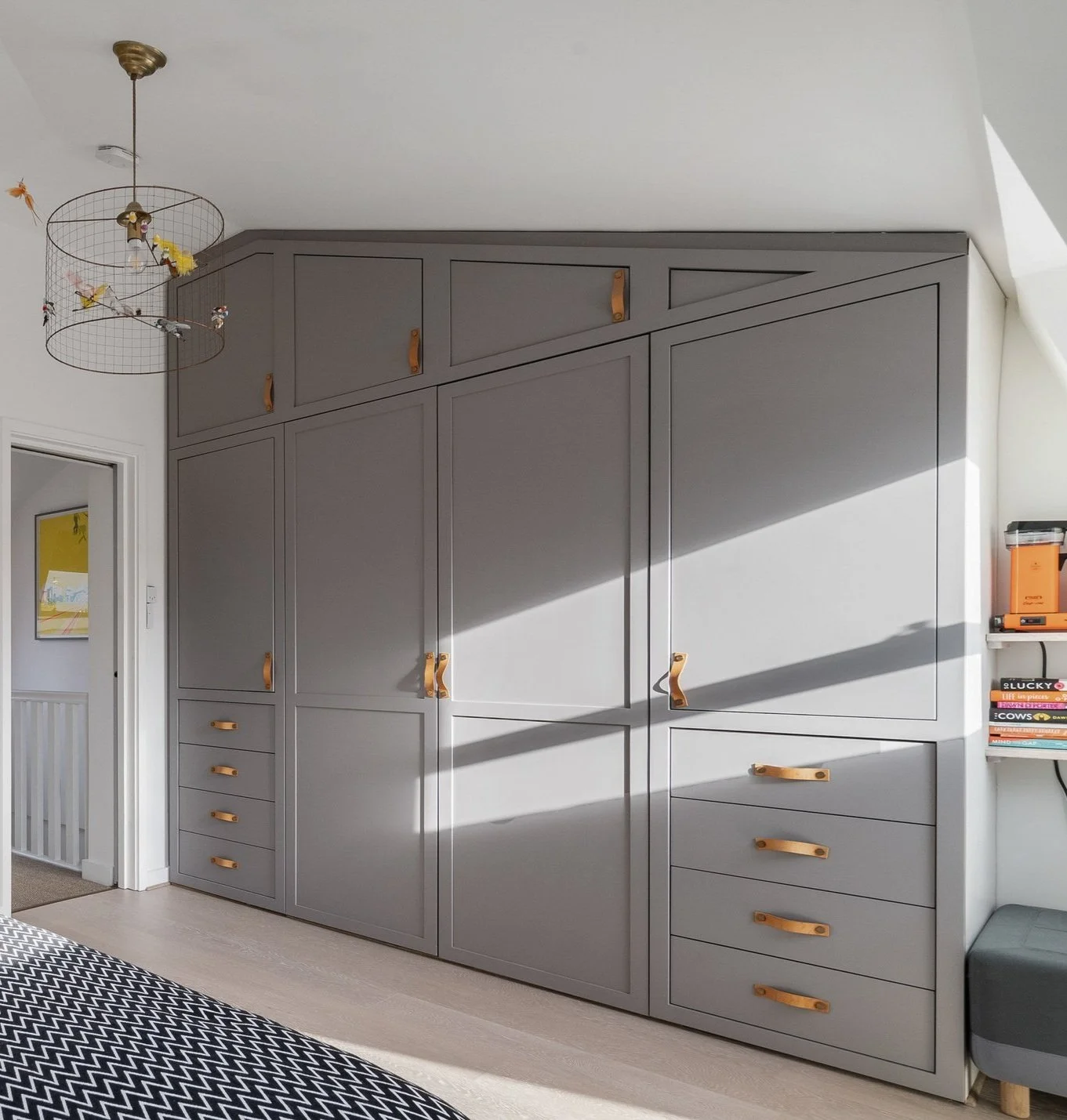 Modern bedroom with built-in gray wardrobe featuring leather handles, geometric pattern bedspread, and hanging wire pendant light. Sunlight casts shadows on the wardrobe. Books and an orange object sit on nearby shelves.