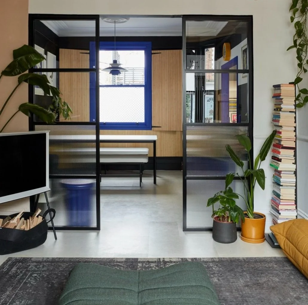 Modern living room with glass partition, green cushion, stack of books, potted plants, and a visible bench in a wooden-finished room.