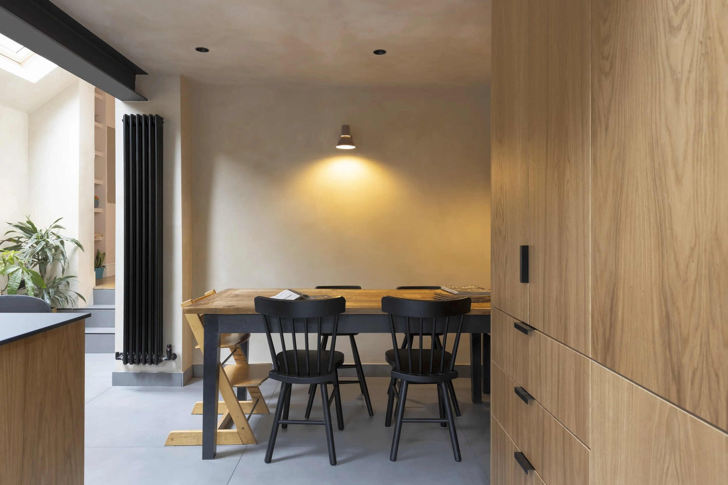 Modern dining area with wooden table and black chairs, adjacent to a wall-mounted light fixture and vertical radiator. Light-colored walls and cabinets, plus plants in the corner, contribute to a minimalist decor.