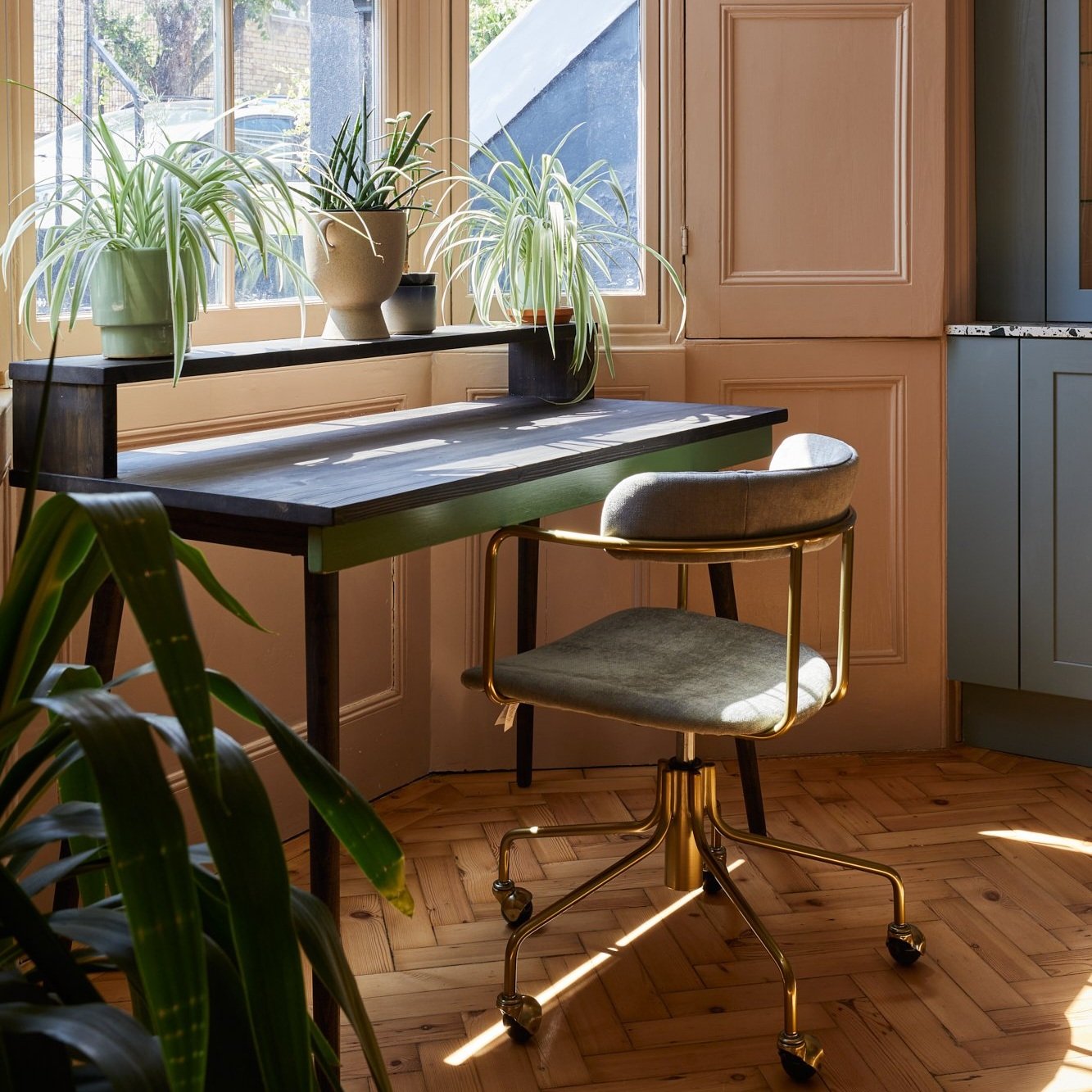 Home office corner with a wooden desk, green upholstered chair with gold frame, and potted plants, next to a sunny window.